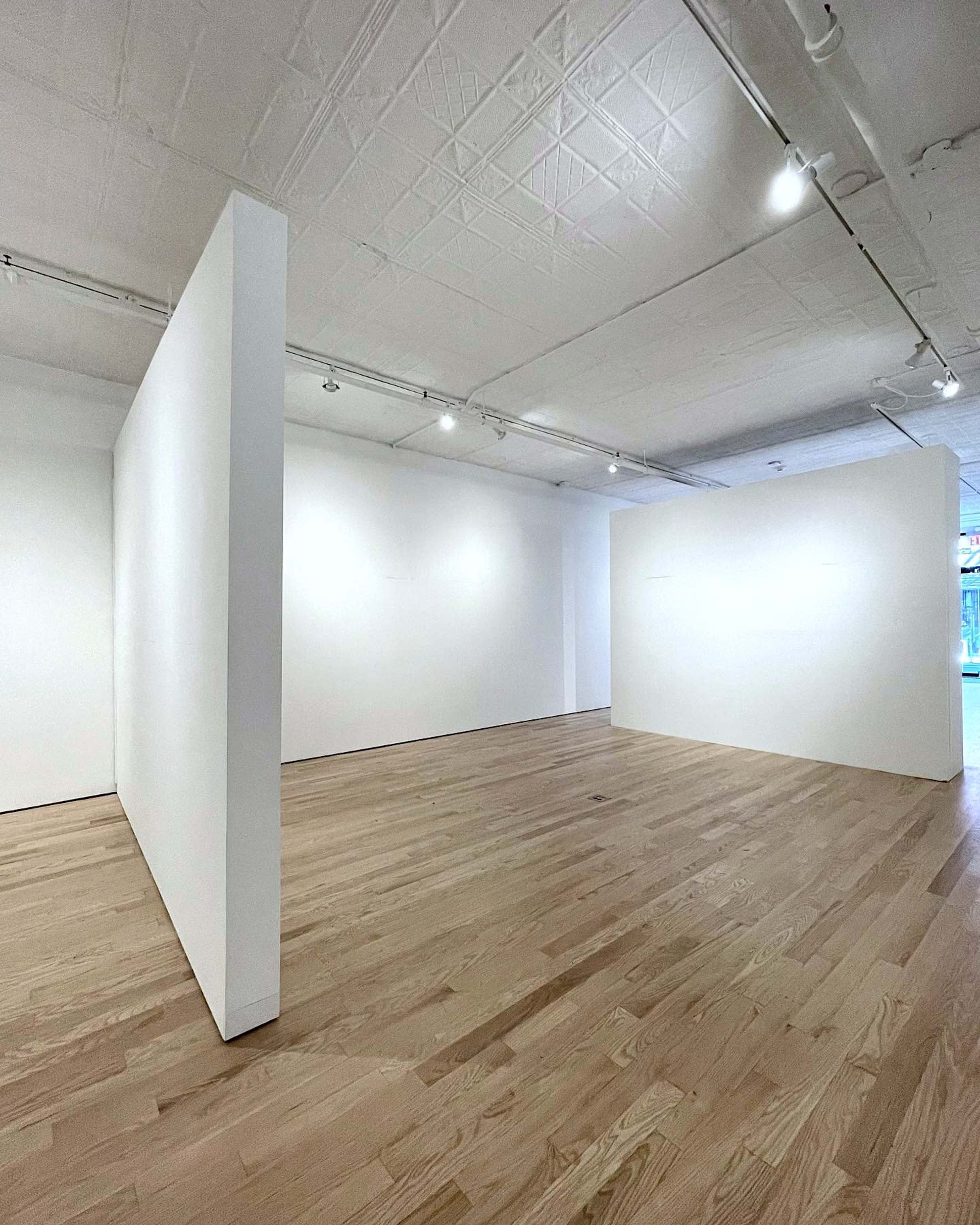 The image shows a spacious, minimalist art gallery with two partially separated white walls and wooden flooring.