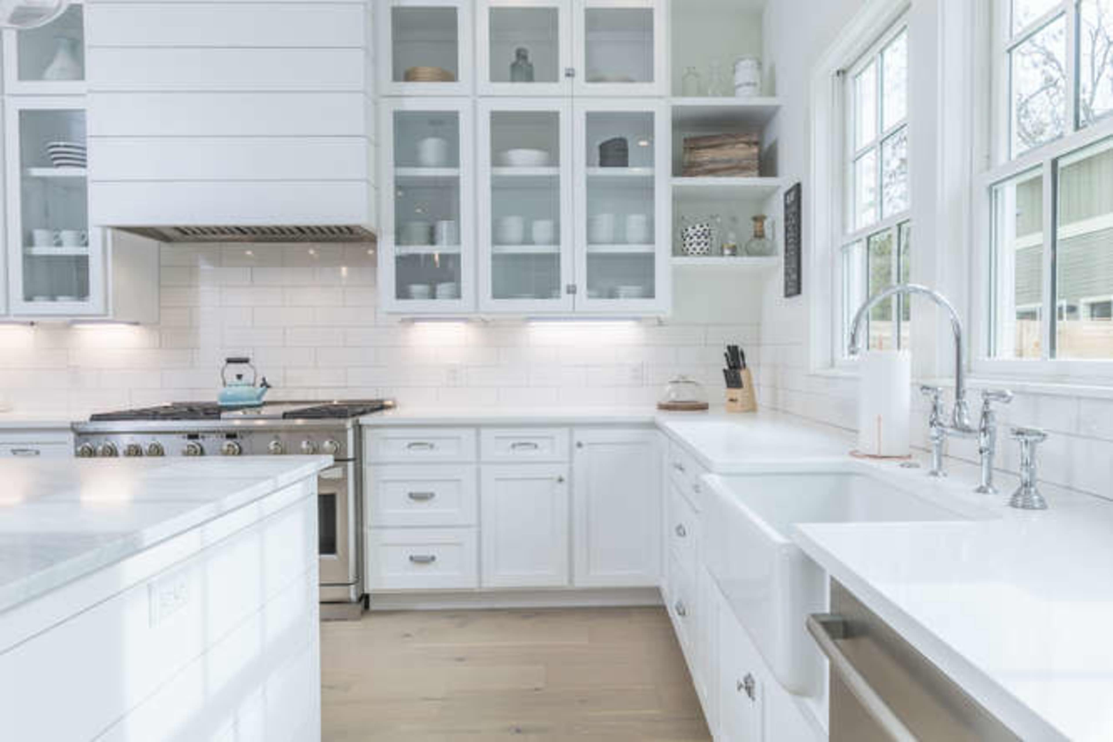 The image shows a modern kitchen featuring white cabinetry, a large sink, a gas stove, and glass-front cabinets displaying dishware.