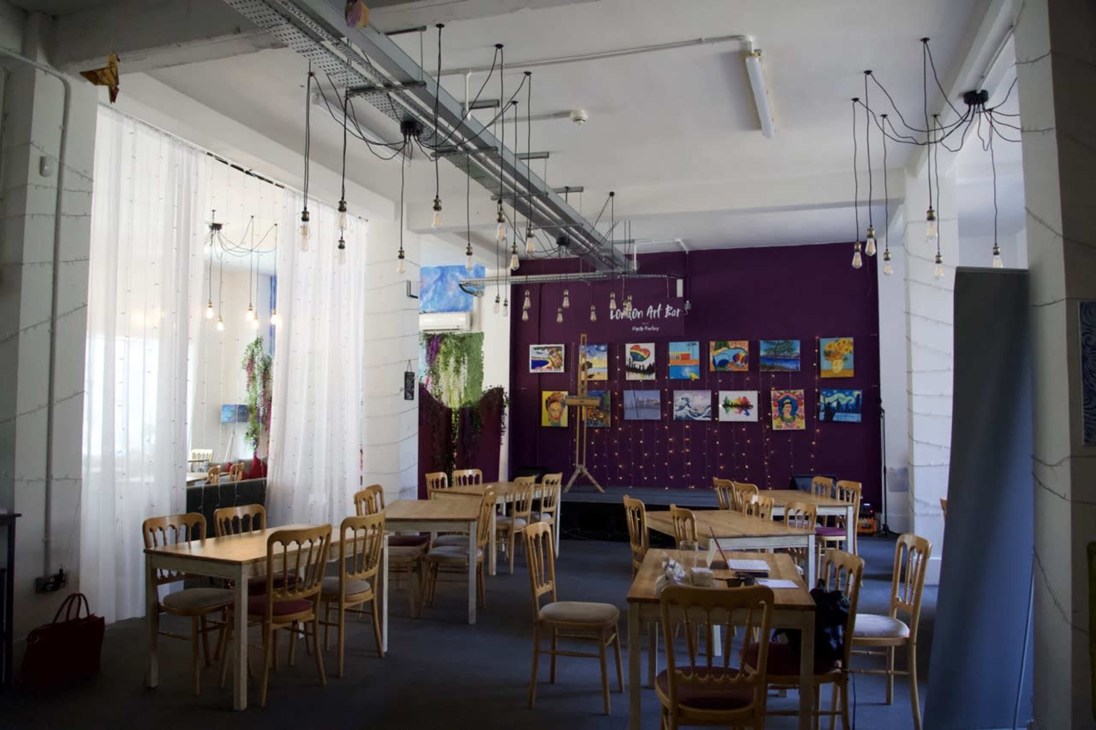 A bright, spacious café features wooden tables and chairs with a purple wall displaying various artworks and hanging light bulbs.