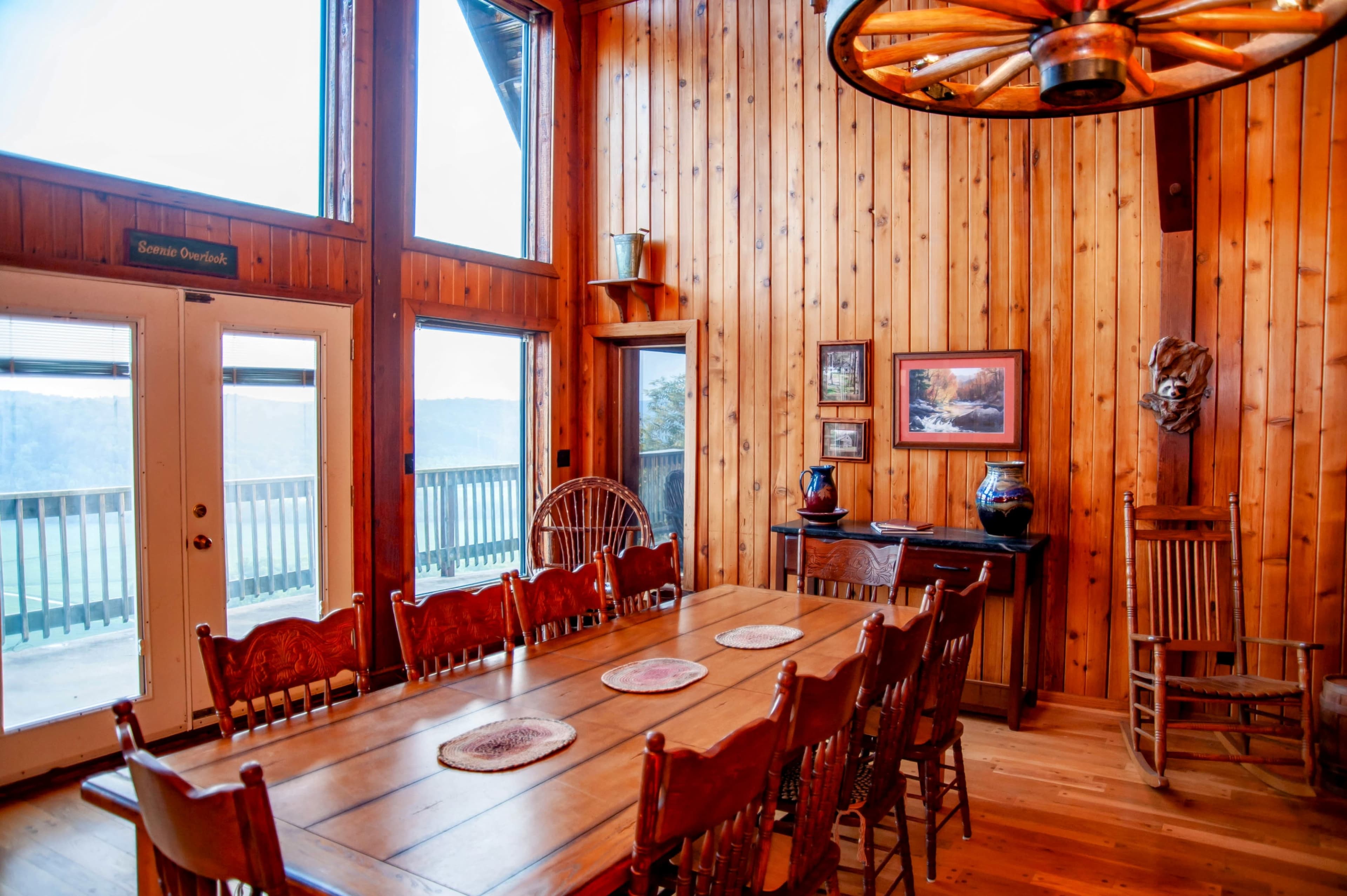 A wooden dining room features a large table surrounded by chairs, with large windows providing a view of the outdoors and wooden paneling on the walls.