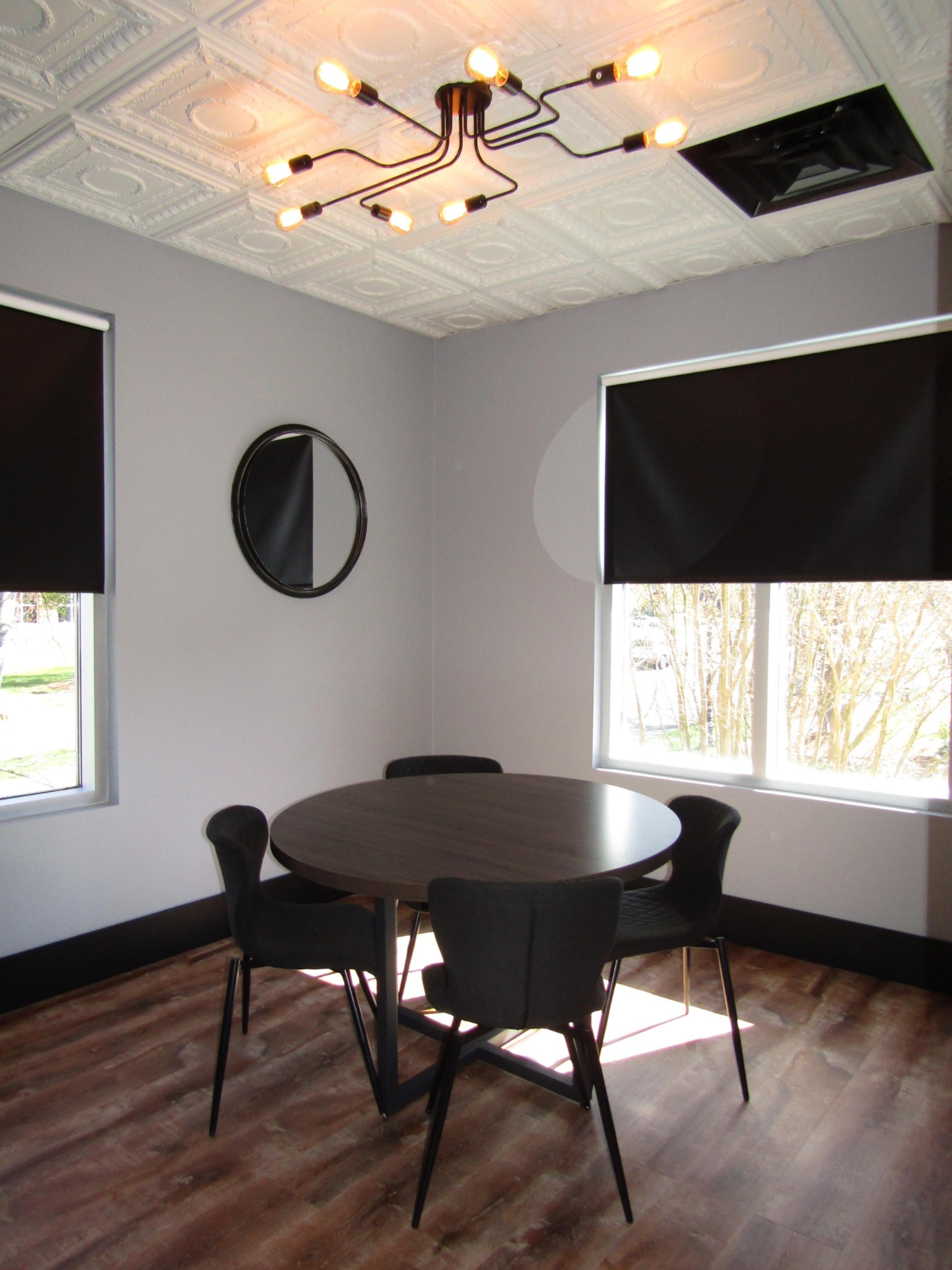 A dining area with a round table and four black chairs under a modern light fixture, featuring gray walls and large windows with black shades.