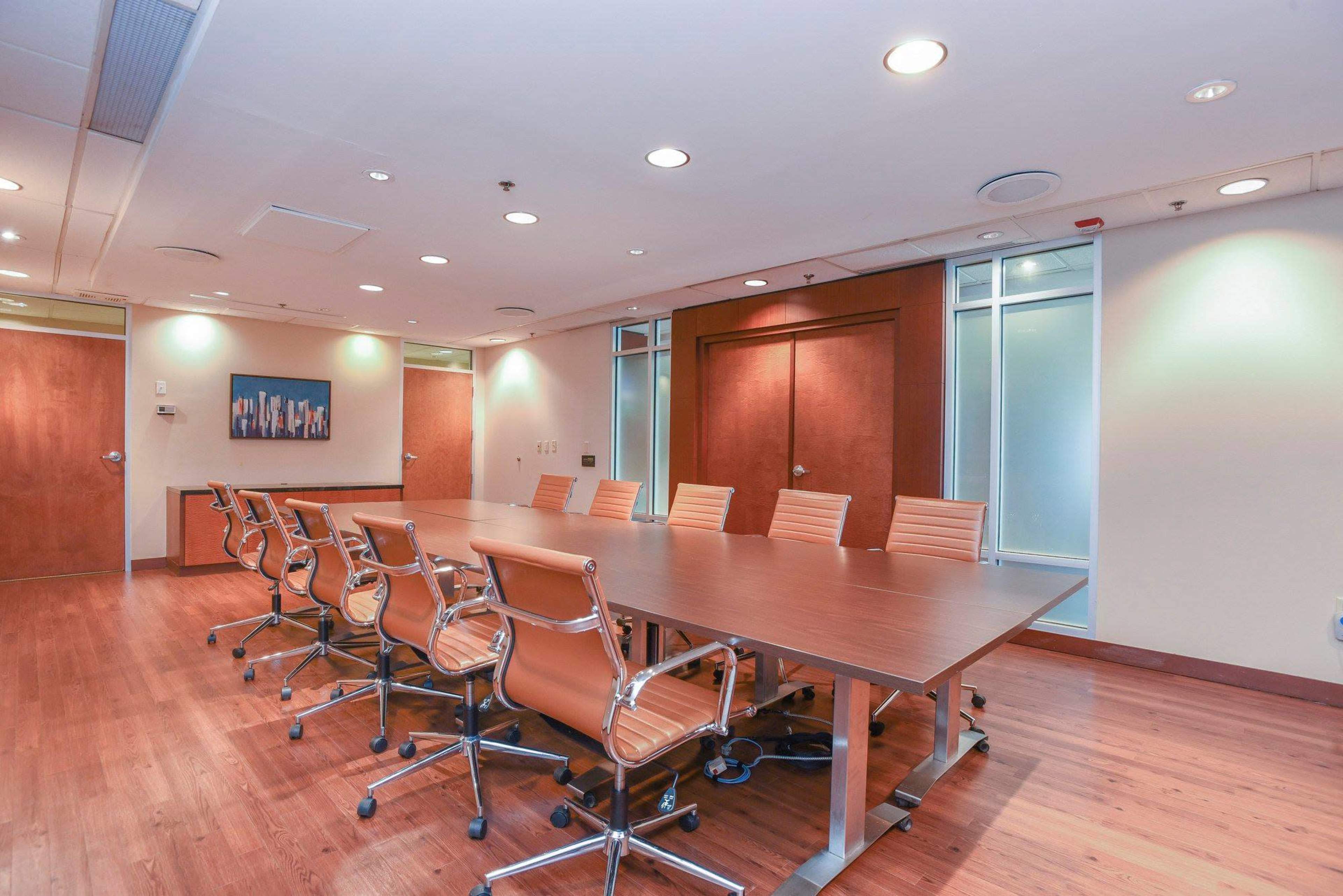 A modern conference room with a long wooden table and eight orange chairs arranged around it, illuminated by overhead lights.