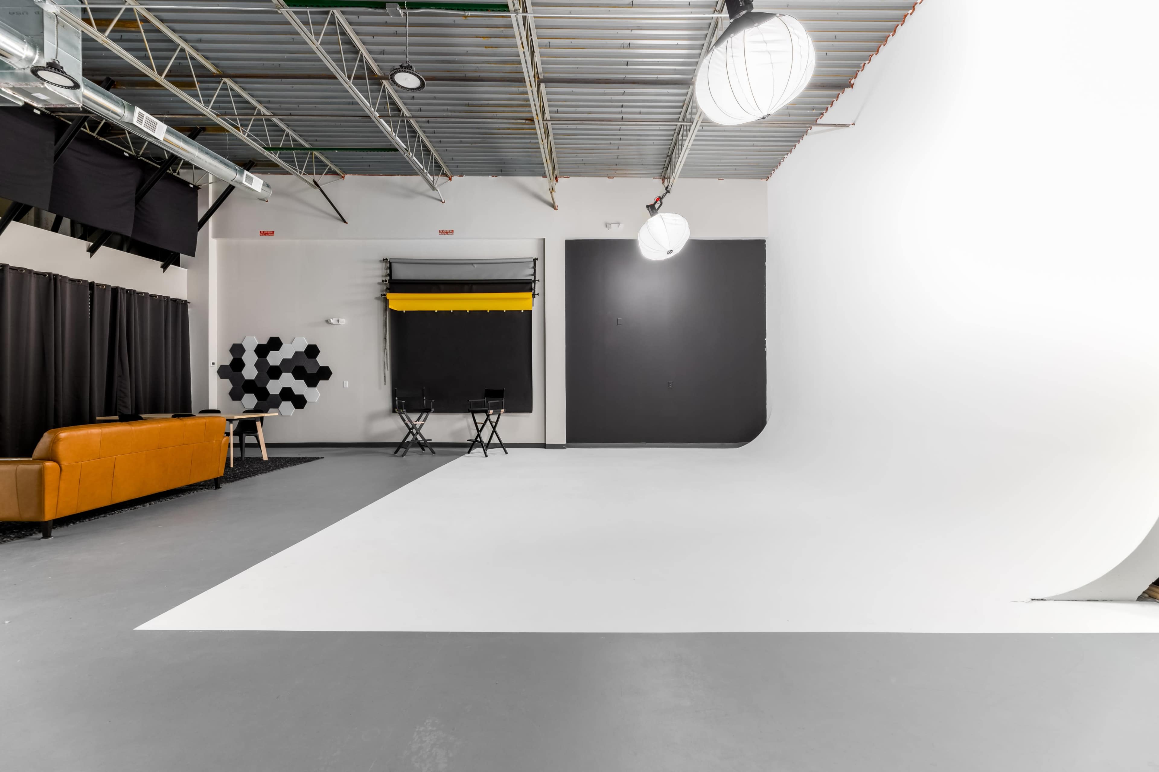The image shows a spacious photography studio with a white backdrop, two chairs, a brown sofa, and ceiling lights.