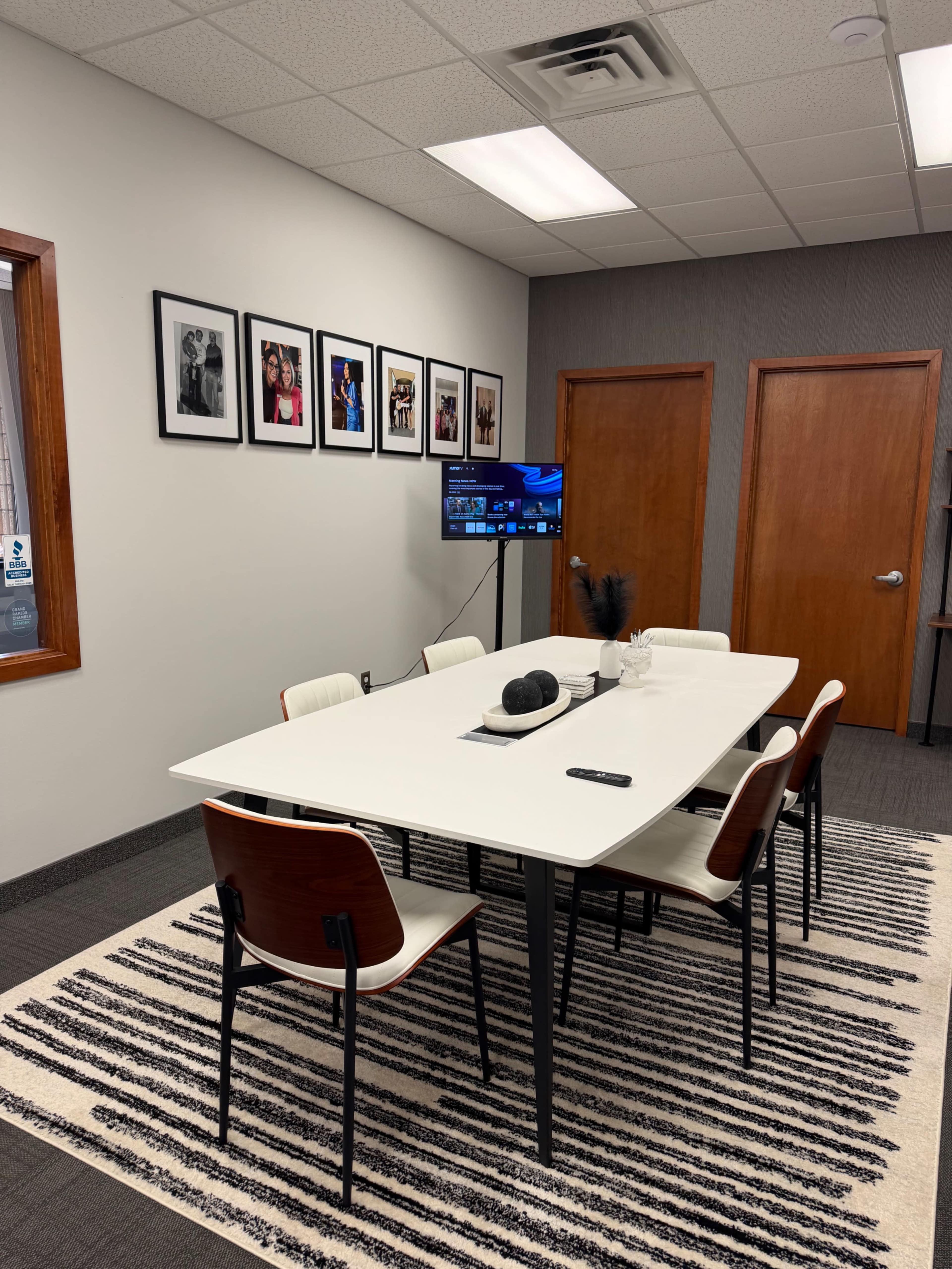 The image depicts a modern conference room with a large white table surrounded by six chairs, a television mounted on the wall, and framed photographs hanging above the table.