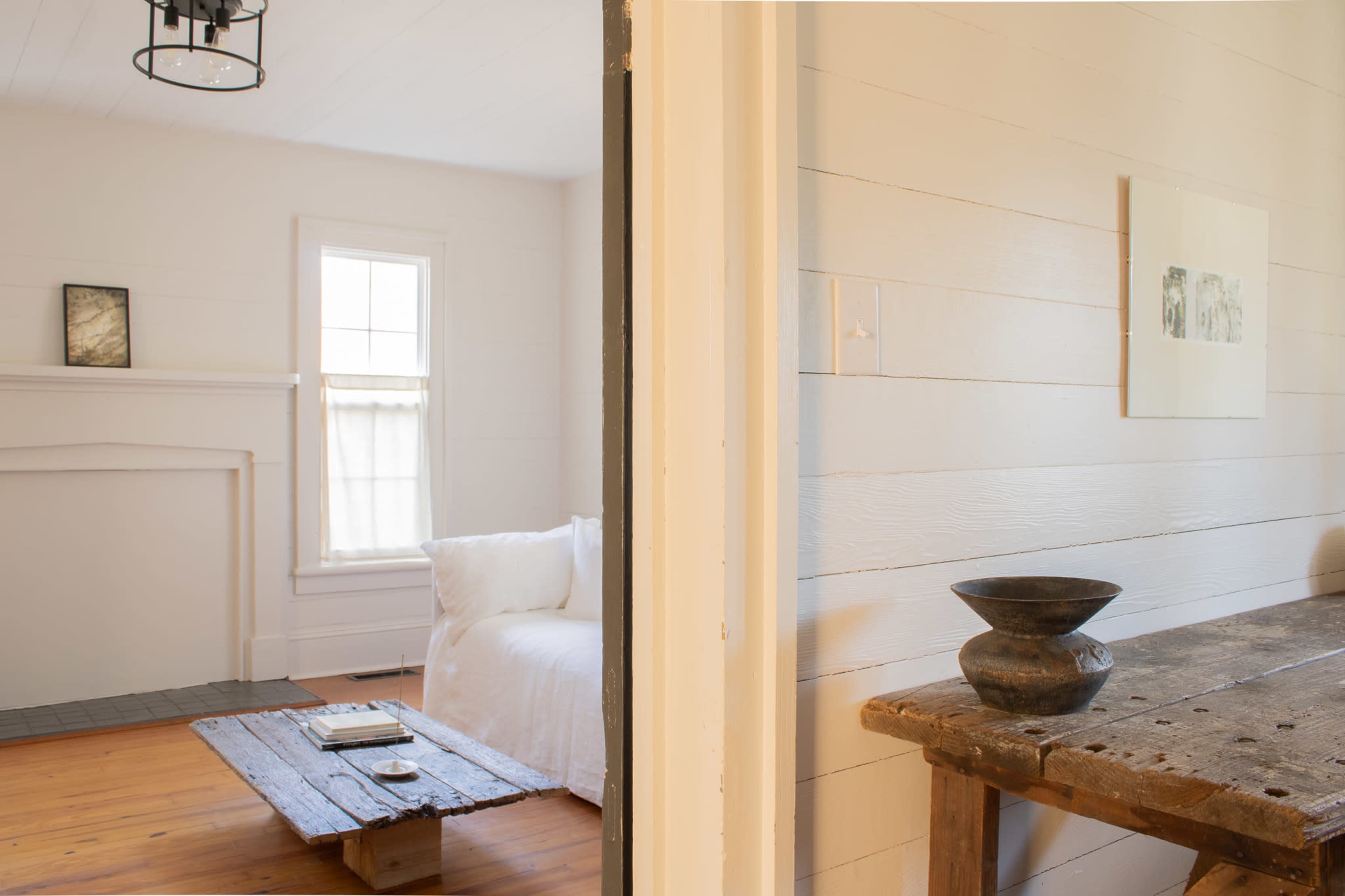 The image shows a bright, minimalist interior with a cozy sitting area featuring a white couch and a wooden table, separated by an open doorway.