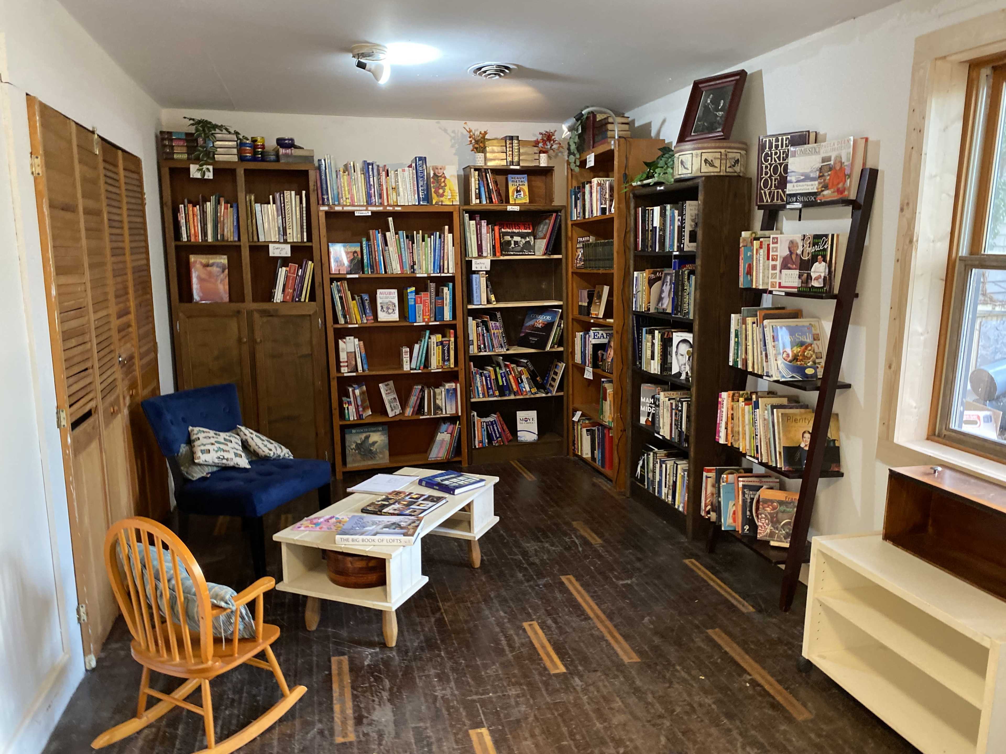 The image shows a cozy reading nook with bookshelves lined with books, a small coffee table, and a blue chair in a room with wooden flooring.