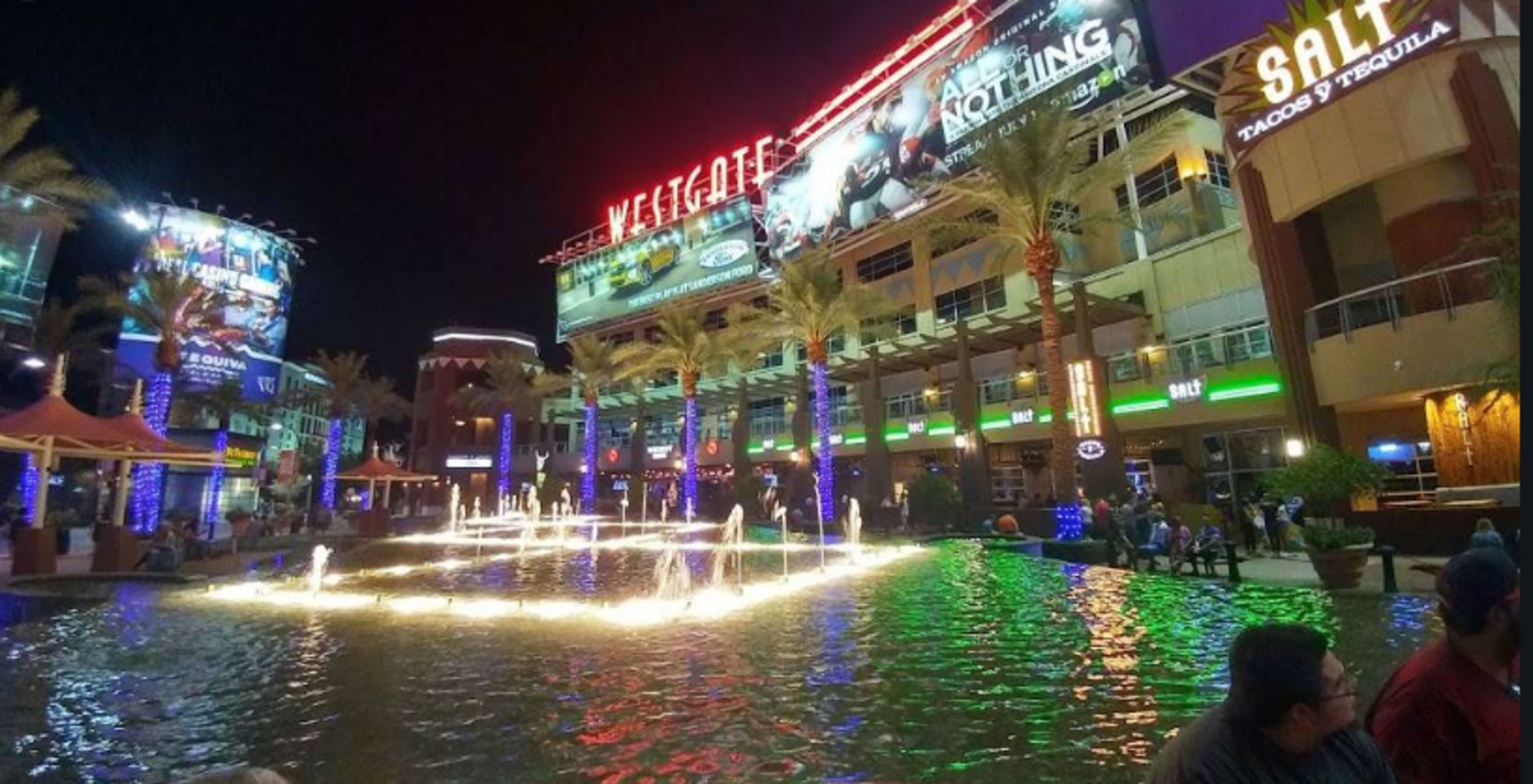 A vibrant shopping and entertainment complex features illuminated fountains and palm trees at night.
