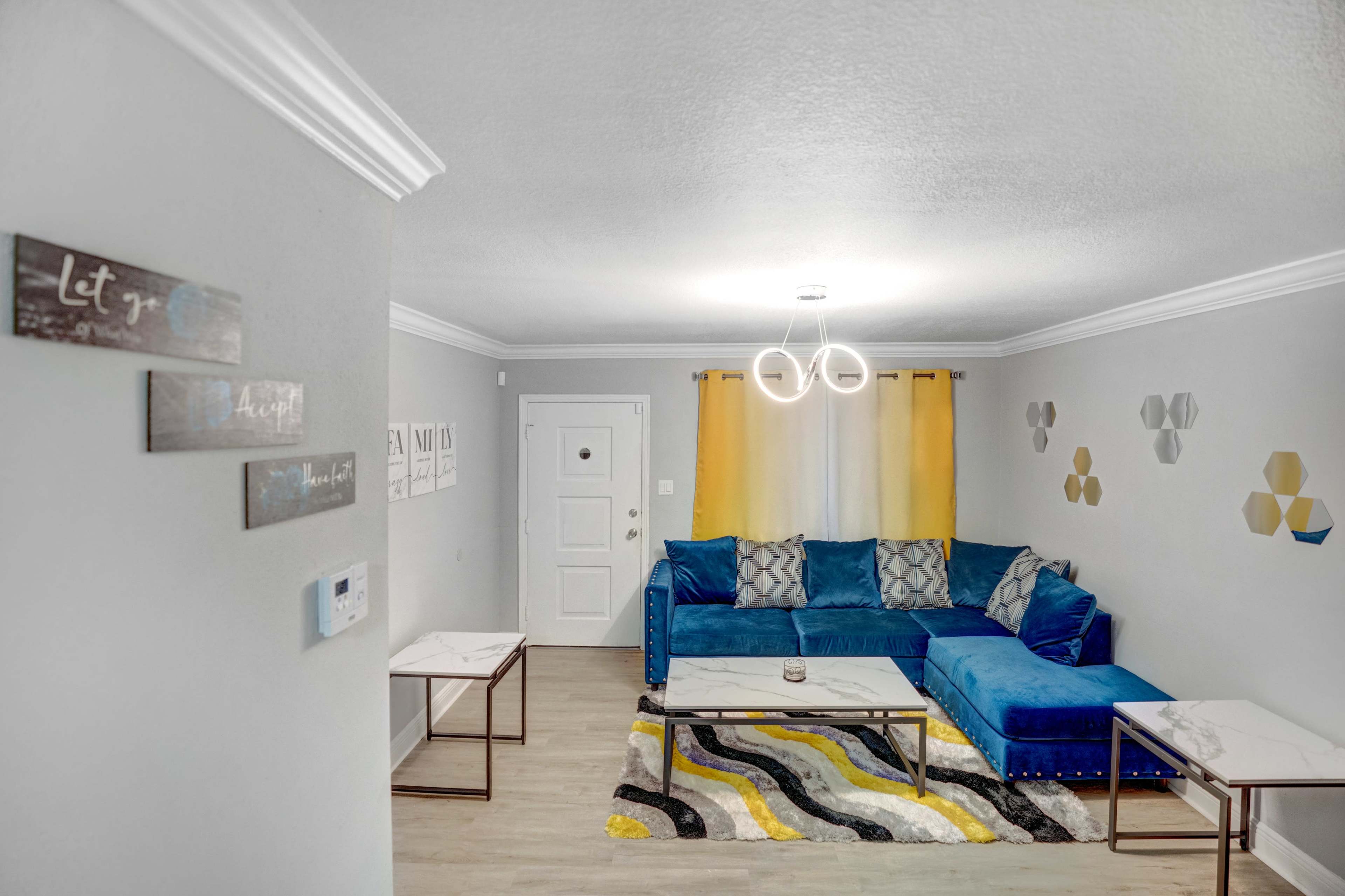 The image shows a modern living room with a blue sectional sofa, two marble-top coffee tables, and yellow curtains beside a white door.