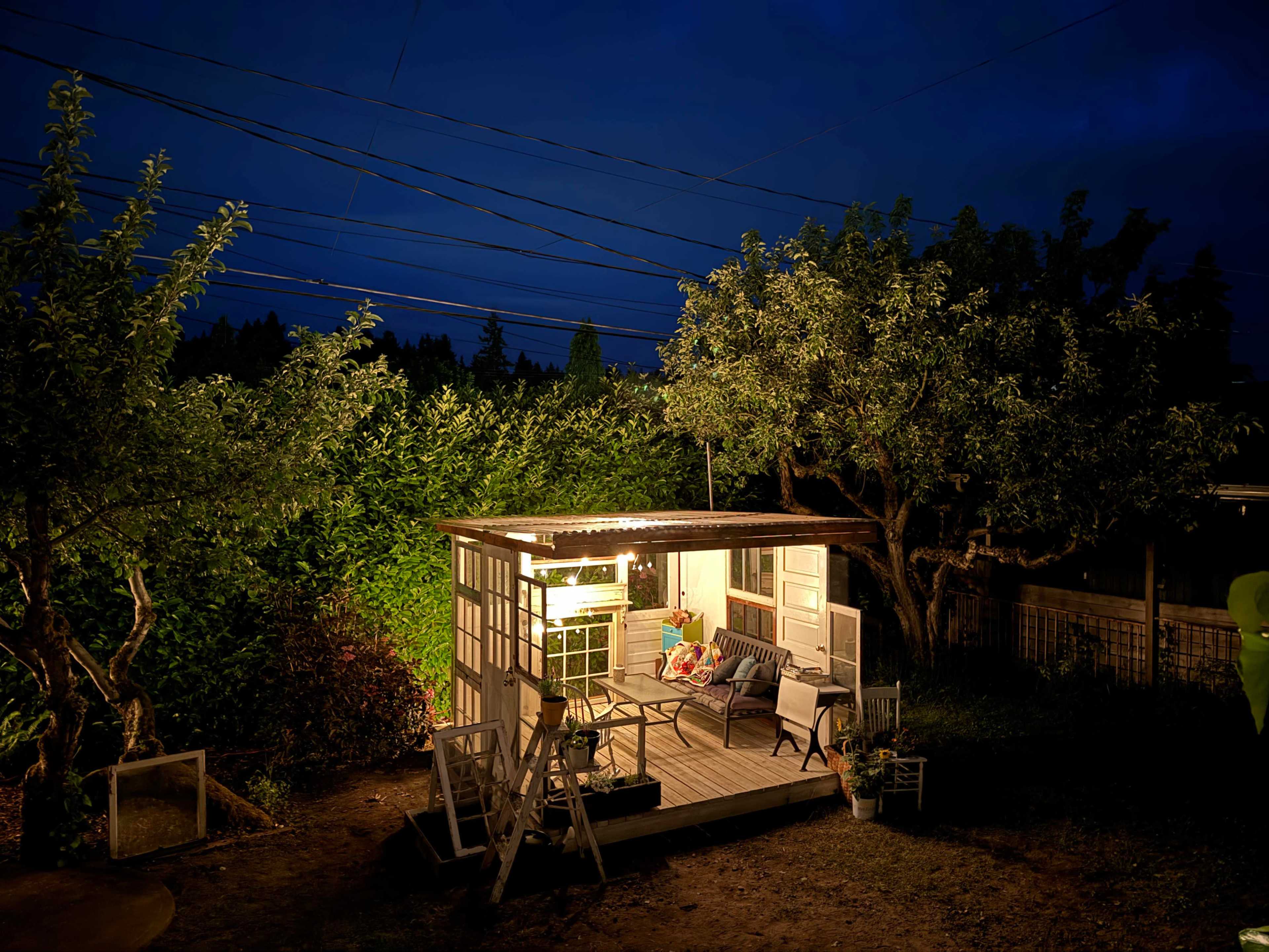 A well-lit shed stands in a dark yard surrounded by trees and power lines.