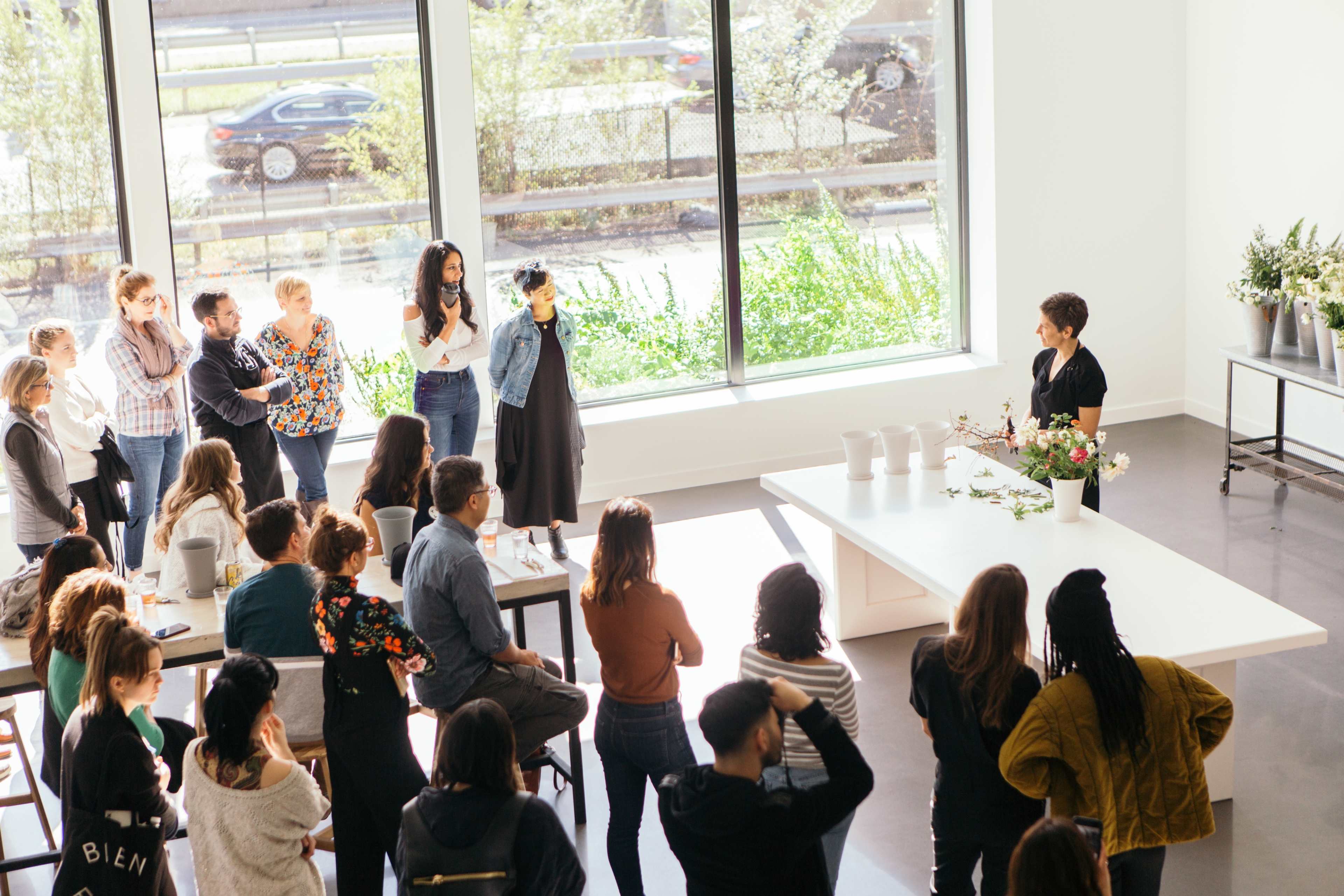 A group of people attentively watches a presentation in a bright, modern space with large windows.