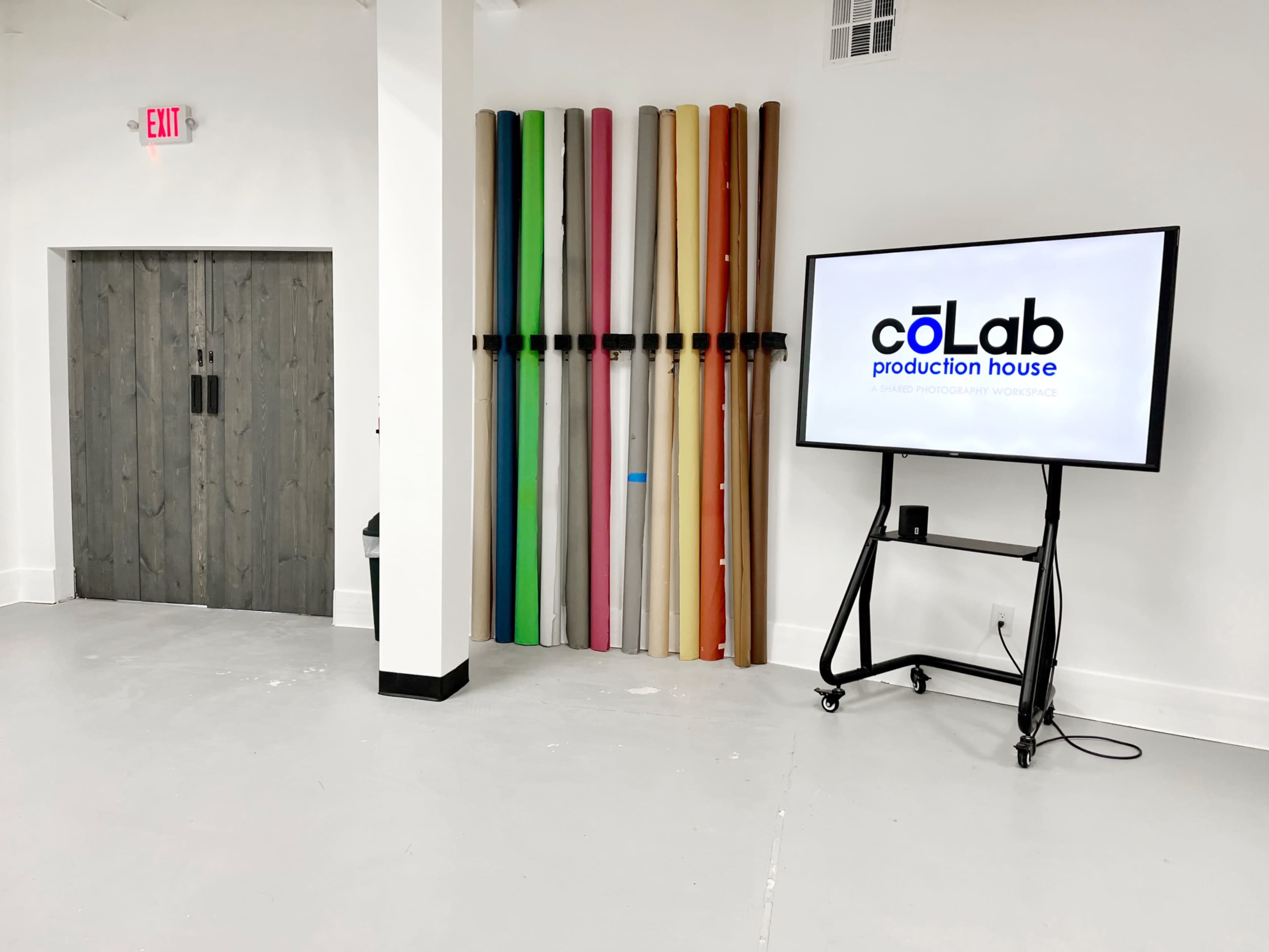 The image shows a spacious room with a large screen displaying the logo of "coLab production house" next to a colorful arrangement of backdrop rolls mounted on the wall.