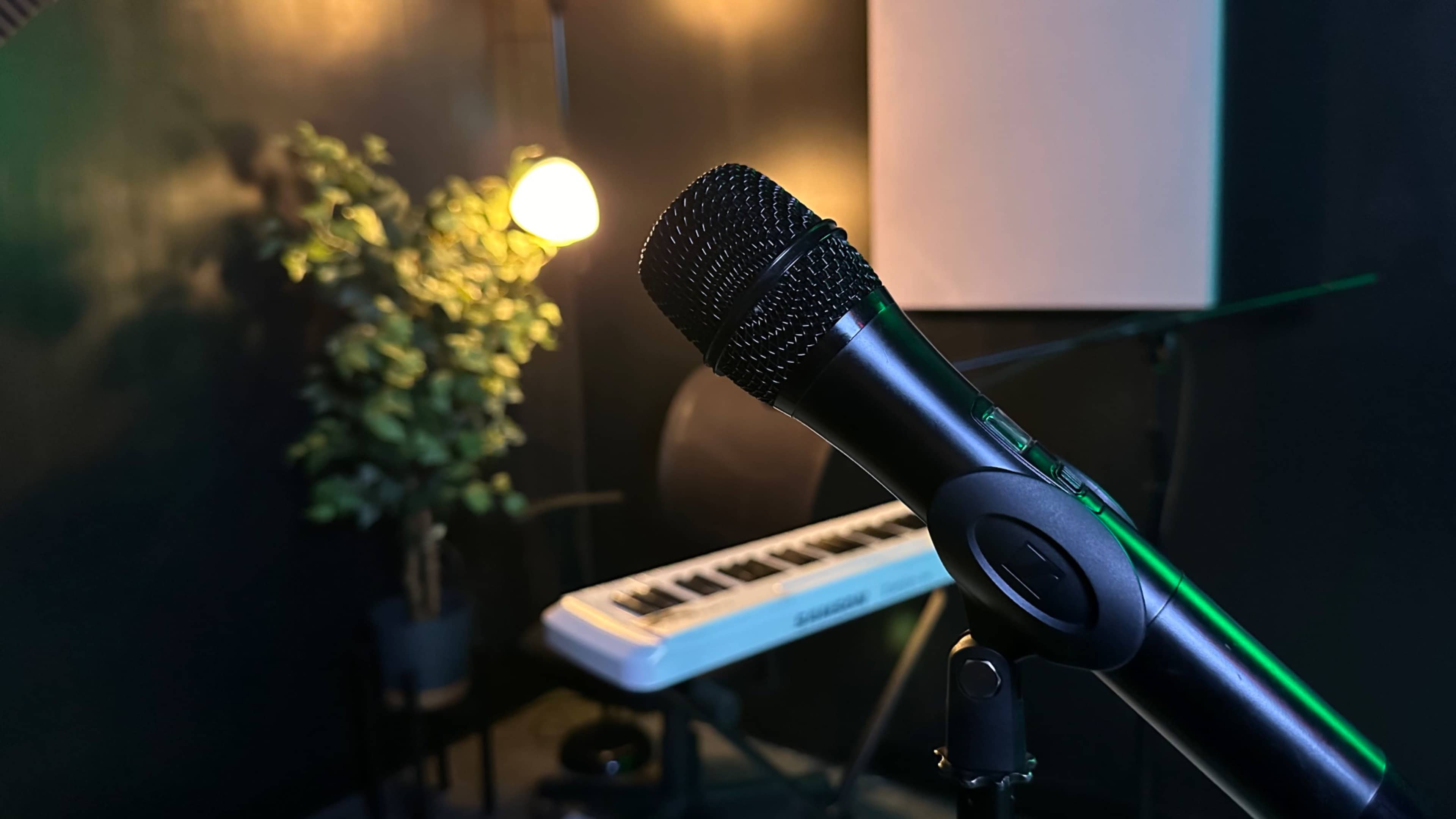 A microphone is in the foreground with a keyboard and a lamp in a dimly lit room featuring a potted plant in the background.