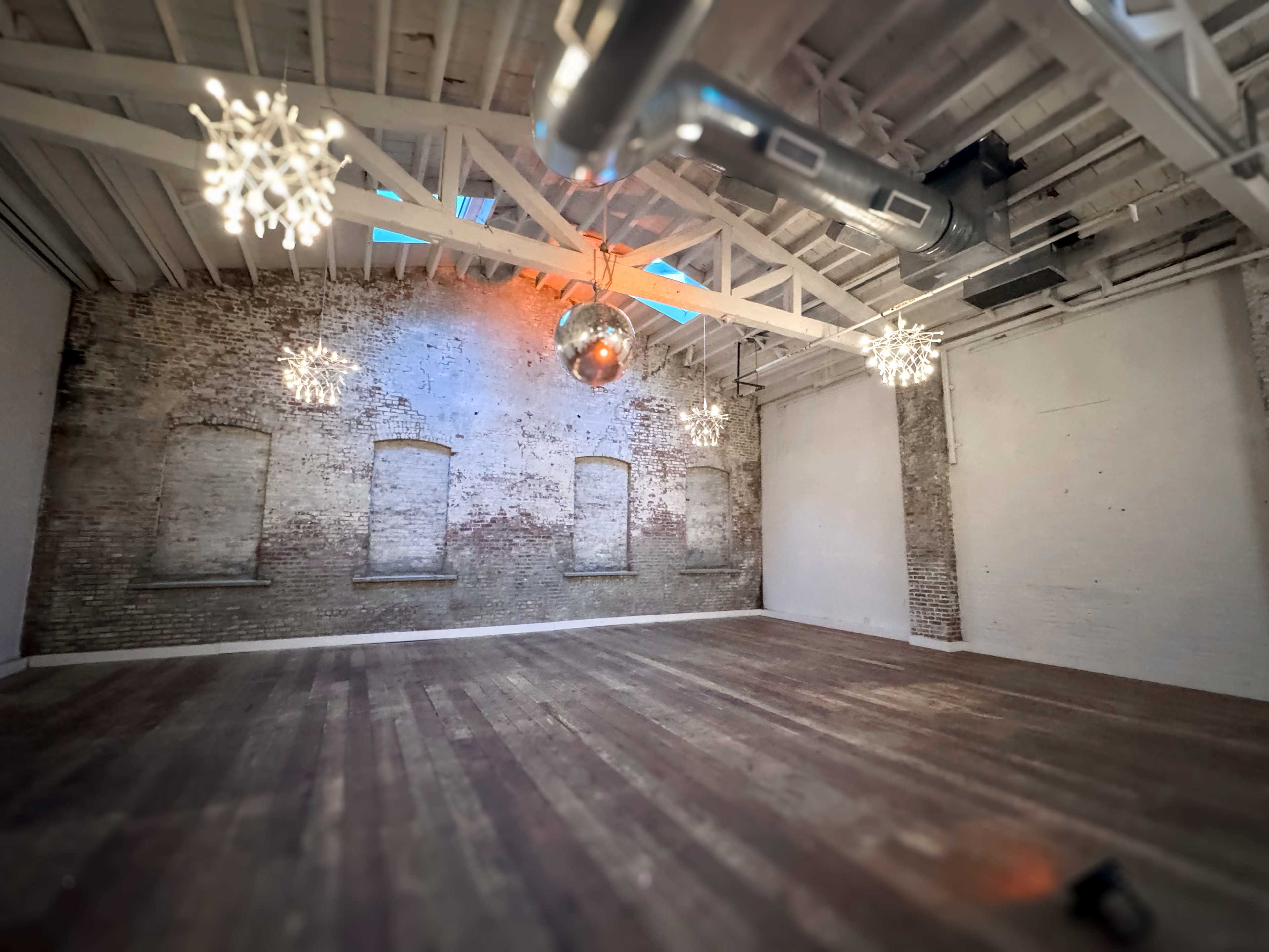 The image shows a spacious, empty room with exposed brick walls, wooden flooring, and several decorative light fixtures hanging from the ceiling.