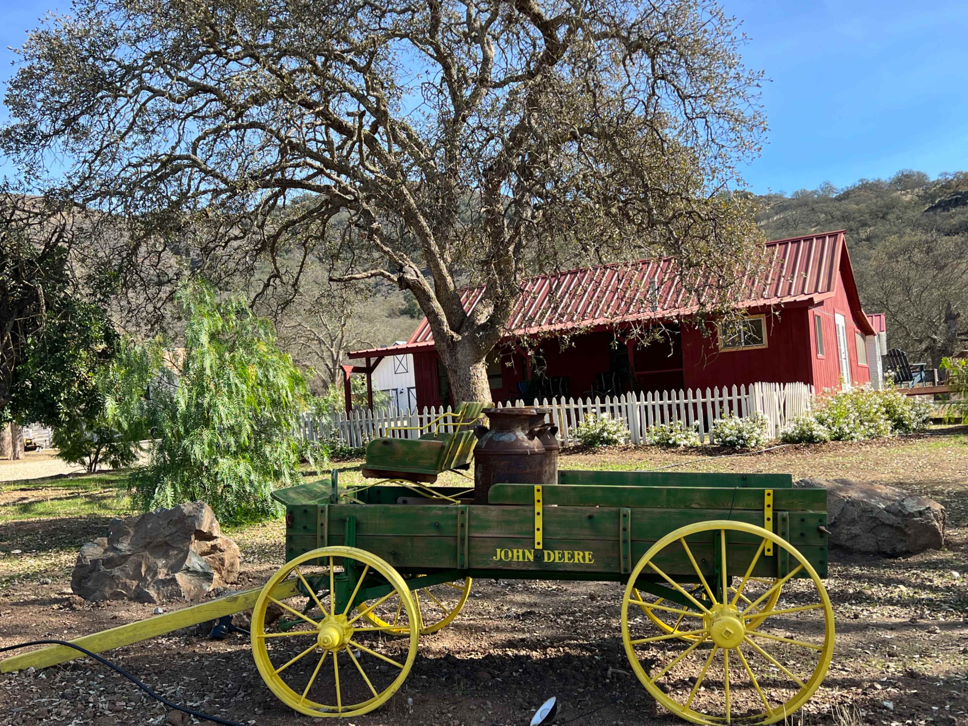 A green wooden wagon marked "JOHN DEERE" sits in front of a red house with a metal roof, surrounded by trees and a white picket fence.