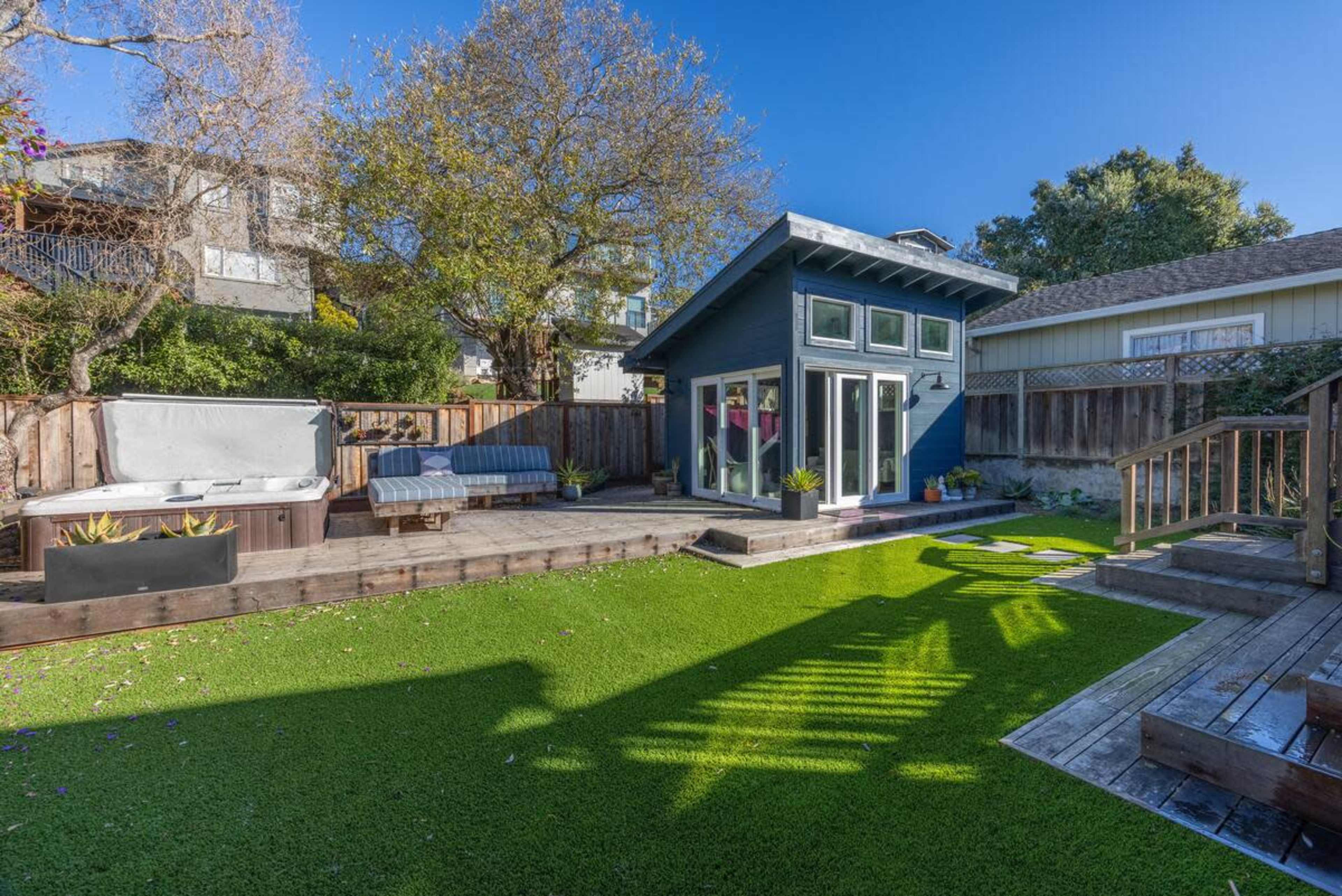 The image showcases a backyard scene featuring a small, modern shed, a wooden deck, artificial grass, and a hot tub surrounded by greenery and trees.
