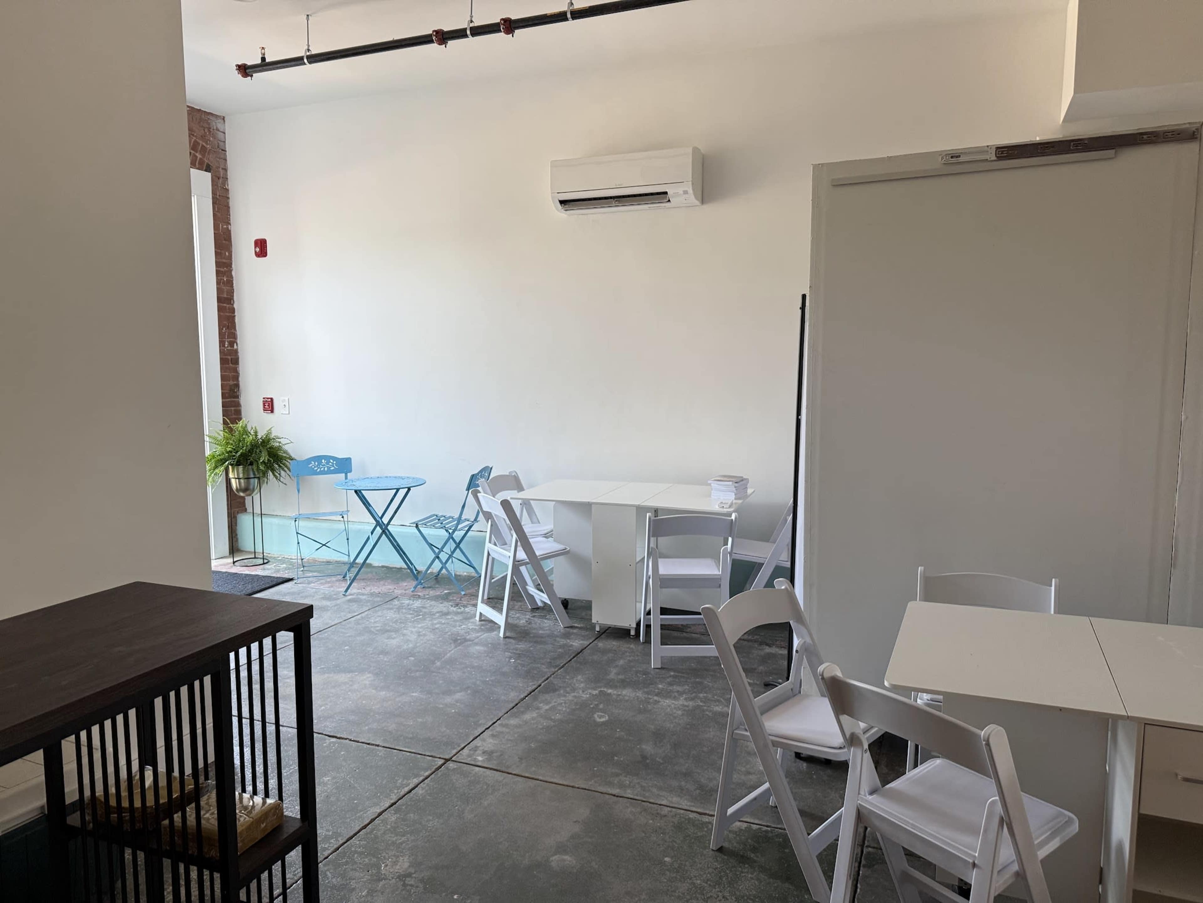 The image shows a spacious, minimally furnished room with white walls, a few tables and chairs, and a small plant near an entrance.