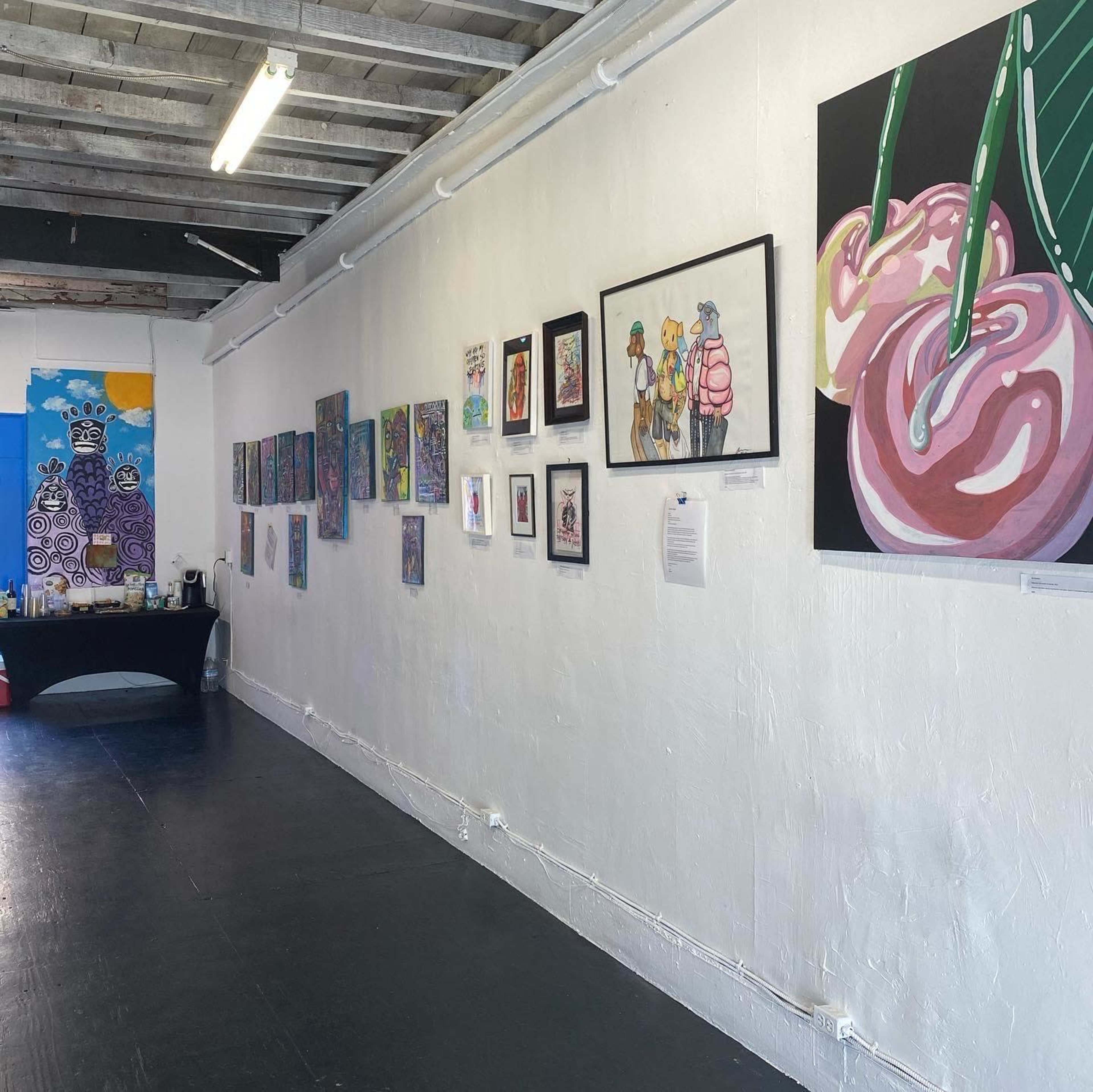The image shows an art gallery featuring various framed artworks displayed on a white wall, with a black table positioned in front.