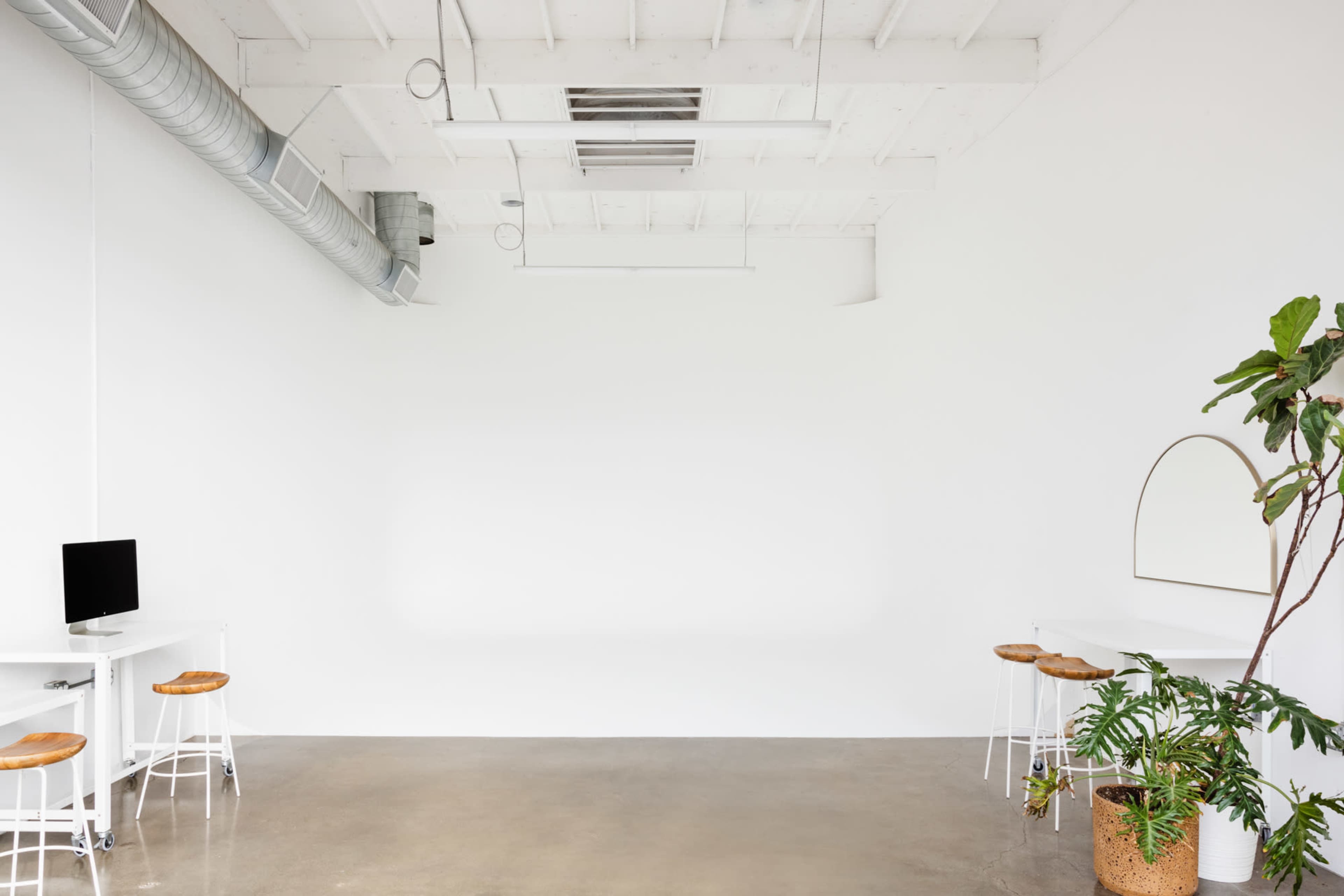 A minimalistic interior space with a polished concrete floor, white walls, and a few pieces of furniture, including a small desk with a computer and two stools.