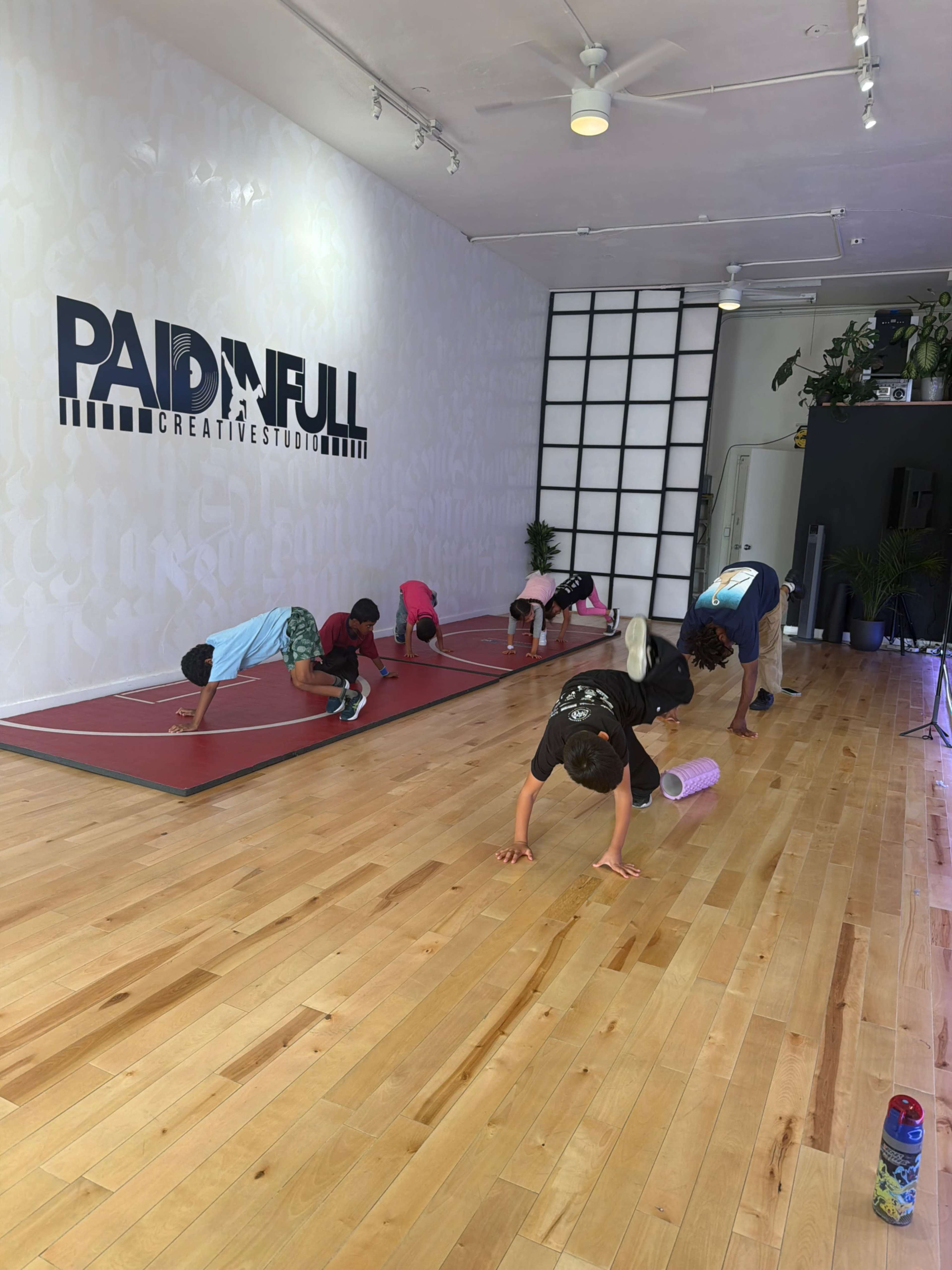Children are practicing gymnastic movements on a wooden floor inside a studio labeled "PAID IN FULL."