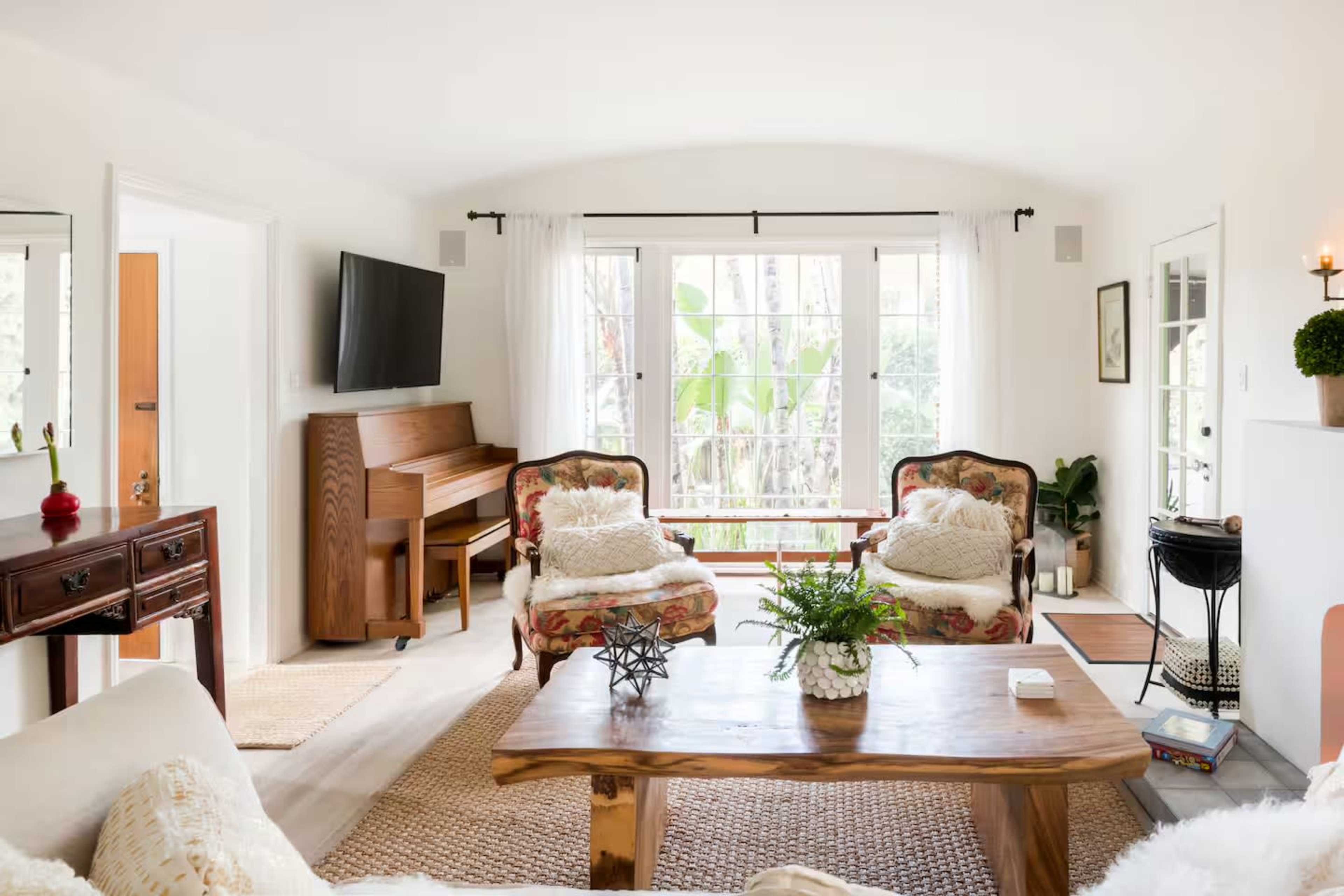 The image shows a bright living room featuring a wooden coffee table, two floral armchairs, a piano, and large windows with leafy outdoor views.