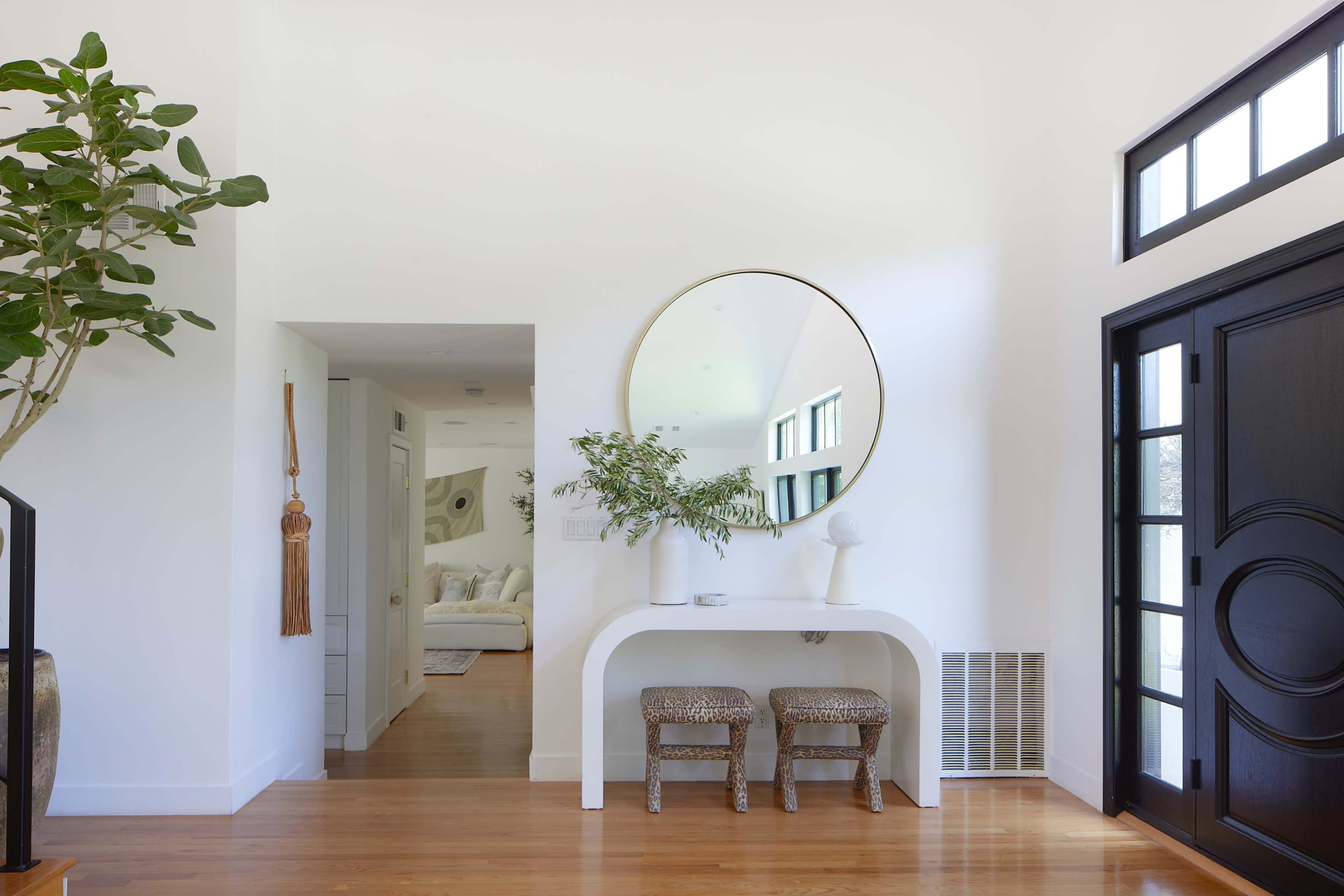 A bright entryway features a round mirror above a white console table with two woven stools and a potted plant.