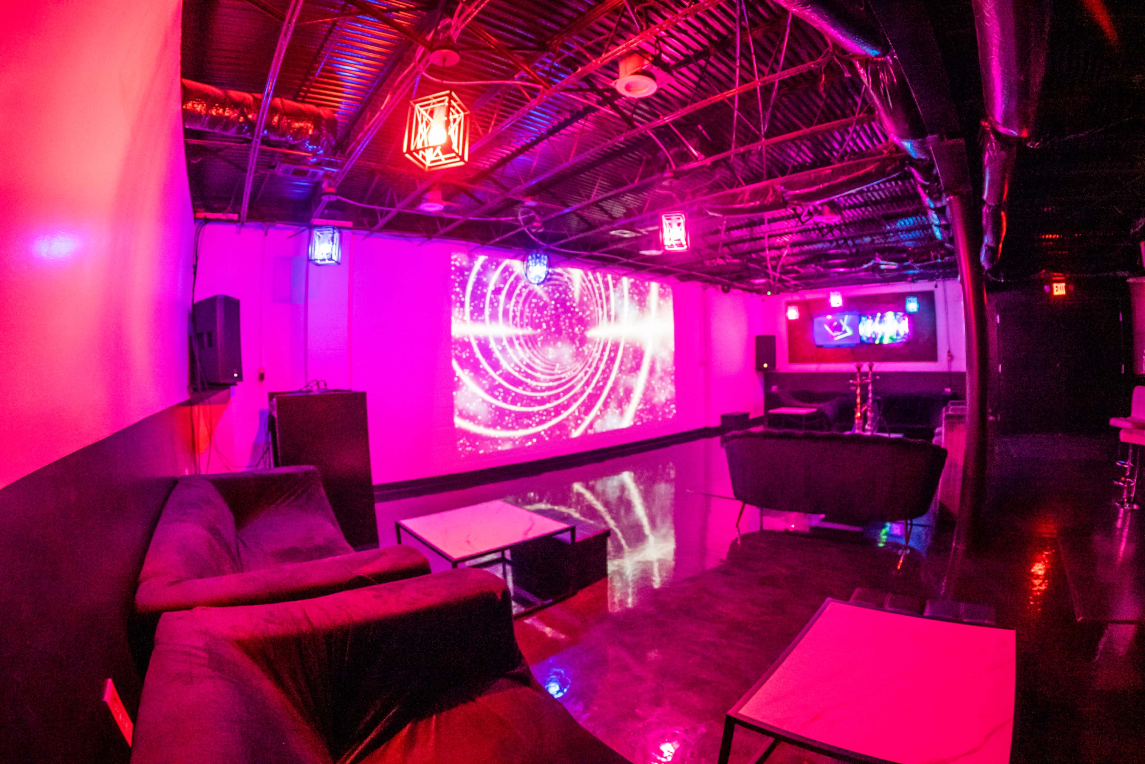 A dimly lit lounge with purple and pink lighting, featuring comfortable seating and an abstract projector display on the wall.