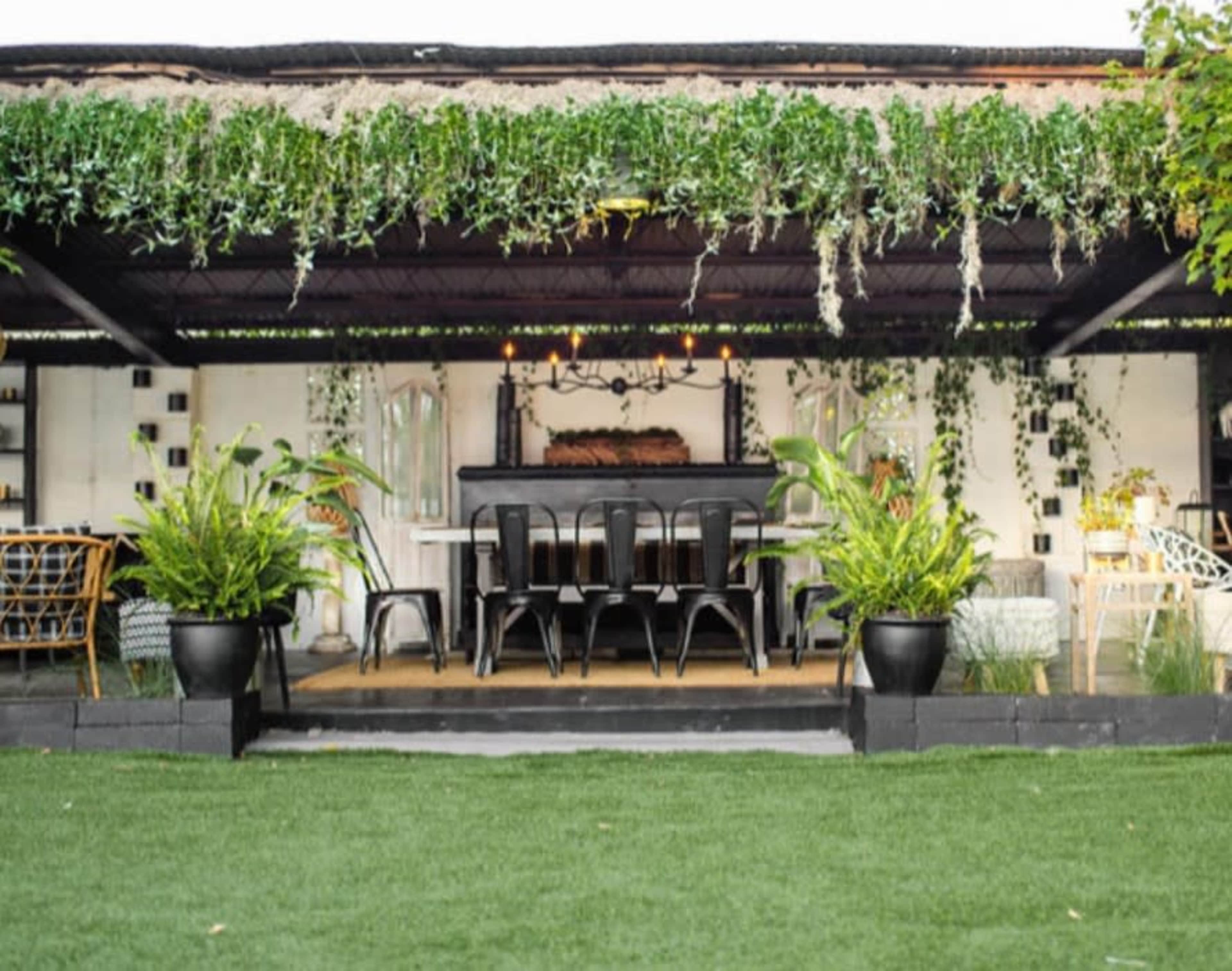 A covered outdoor dining area with a large table surrounded by chairs, adorned with hanging greenery and potted plants.