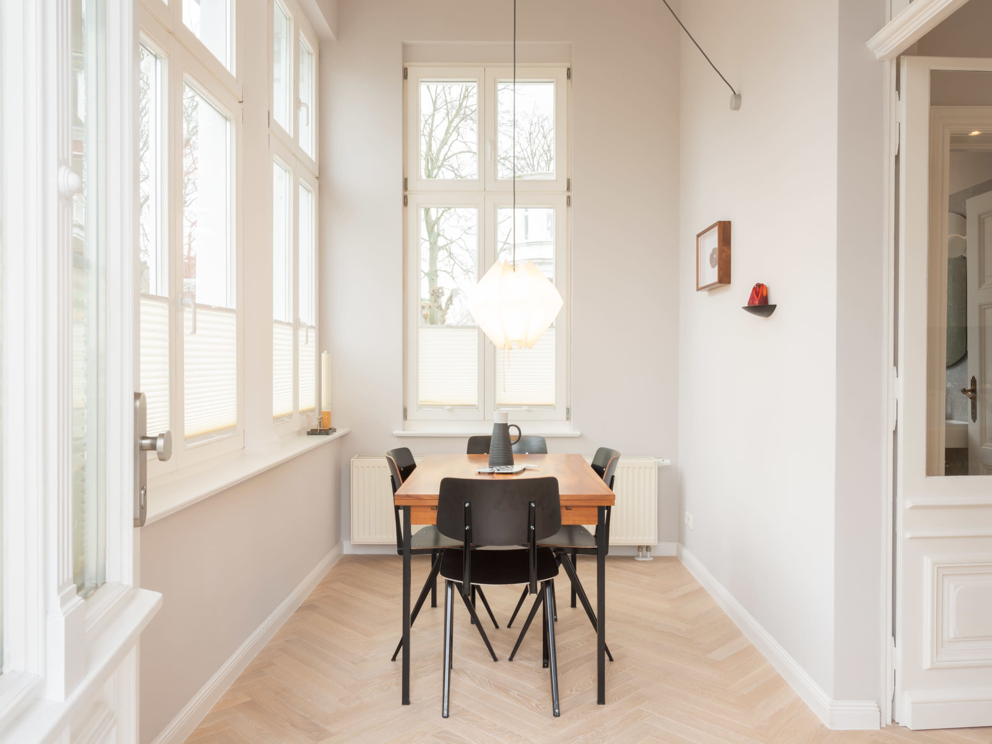 The image shows a bright dining area with a wooden table and four black chairs, illuminated by a pendant light, surrounded by large windows.