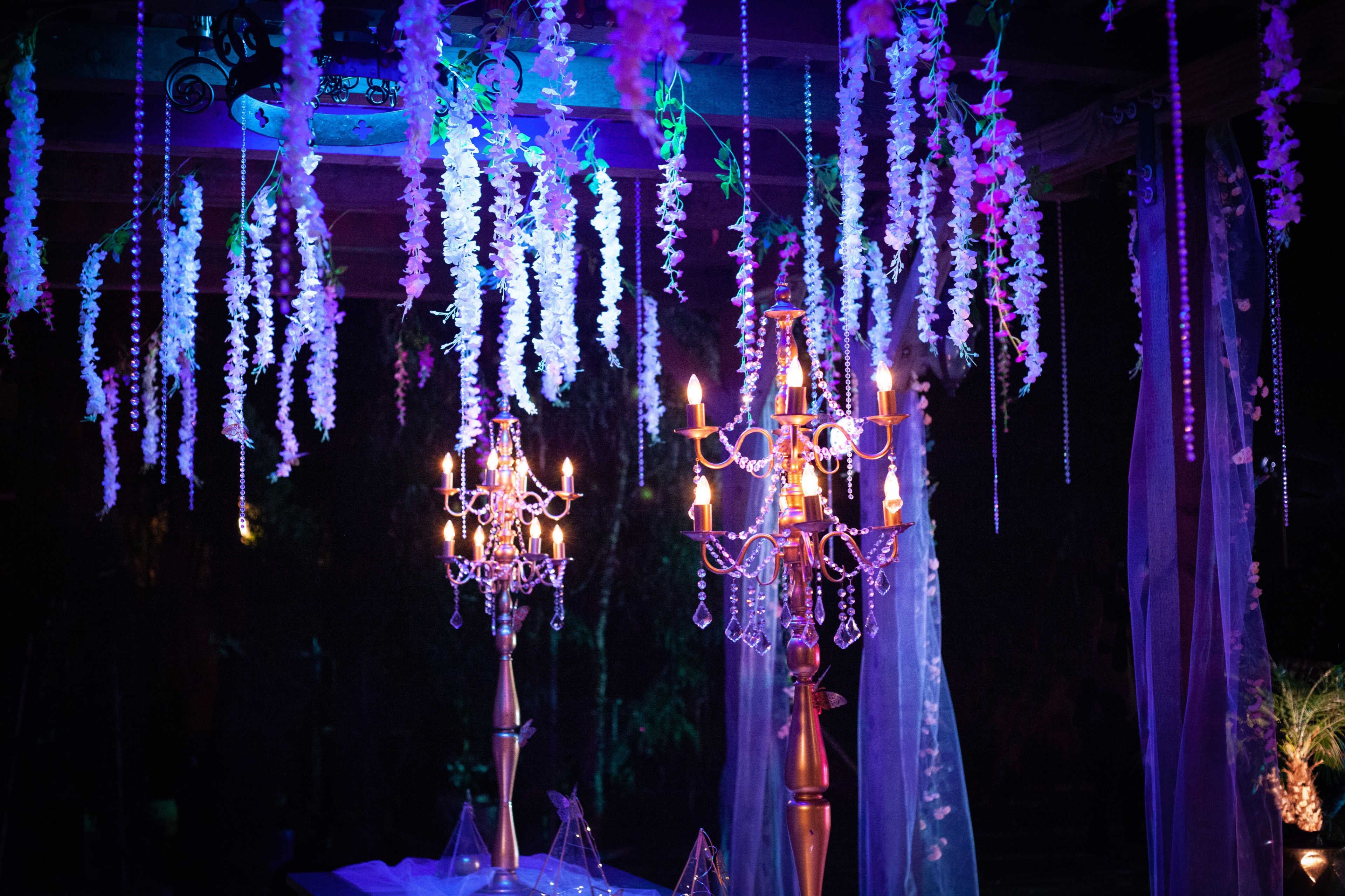 The scene features two elegantly designed chandeliers surrounded by hanging floral decorations and illuminated by colorful lights.