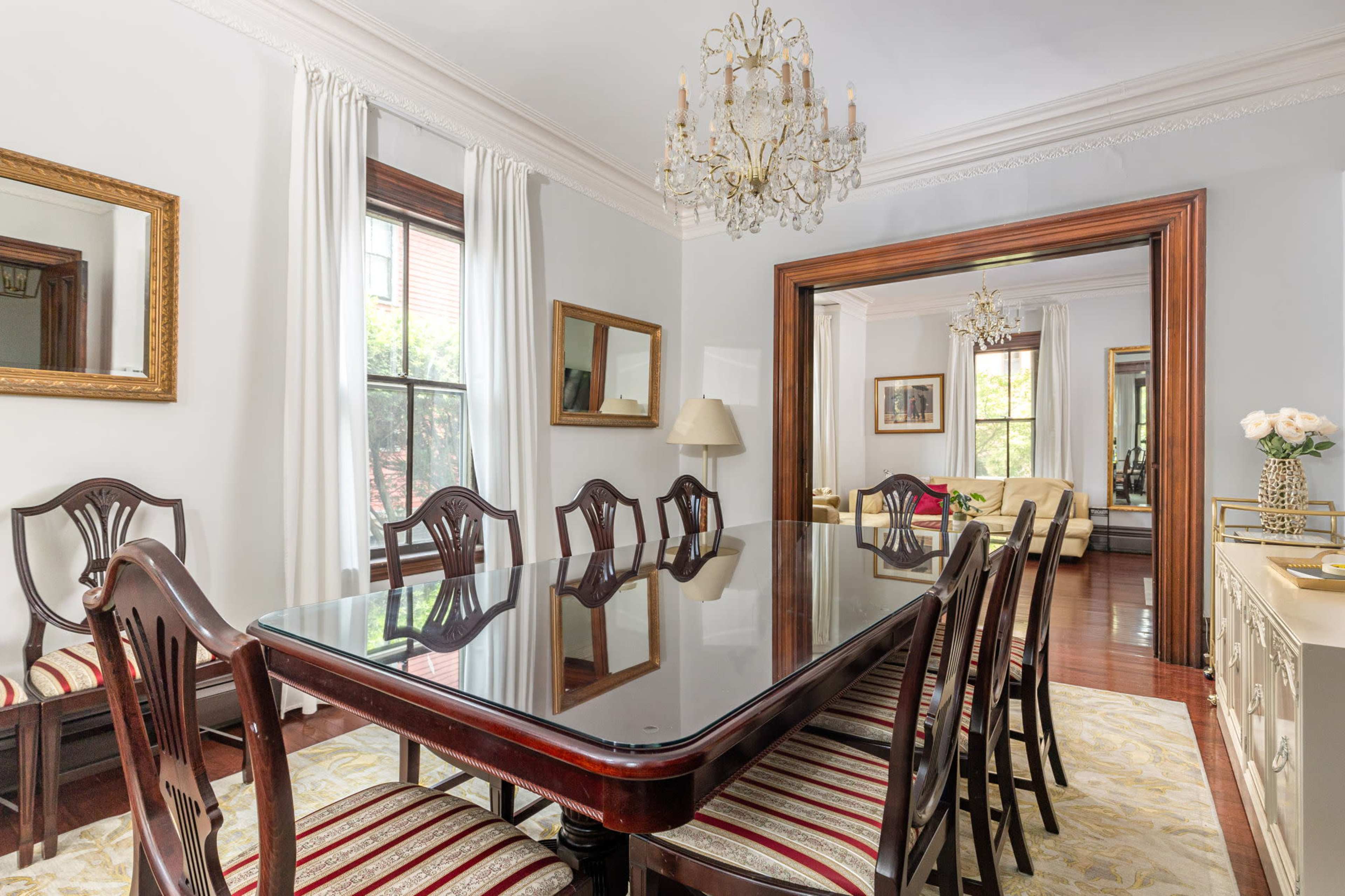 A dining room features a large table surrounded by eight chairs, with a chandelier overhead and windows allowing natural light in.