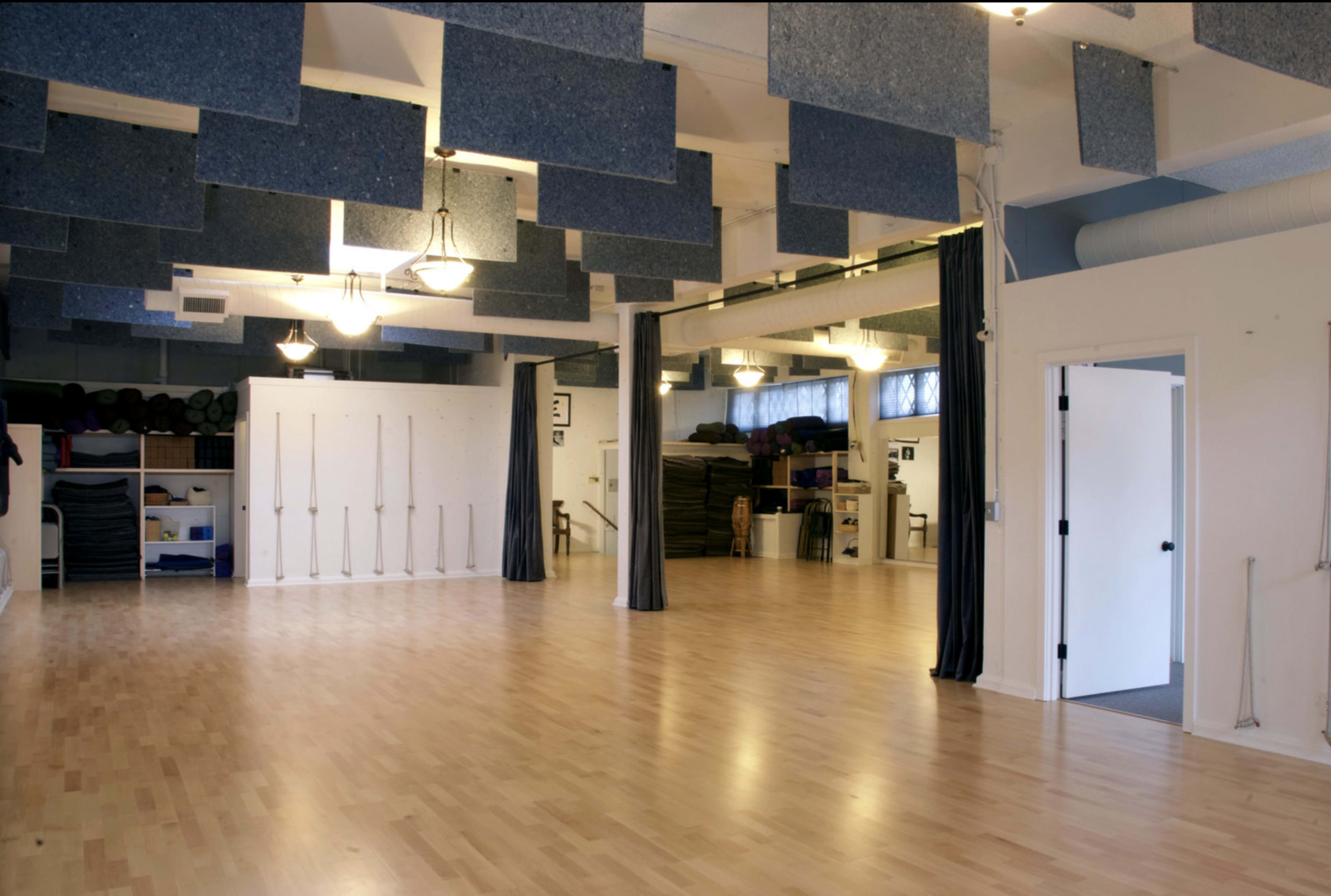 A spacious, well-lit studio with a wooden floor, blue acoustic panels hanging from the ceiling, and curtains dividing different areas.