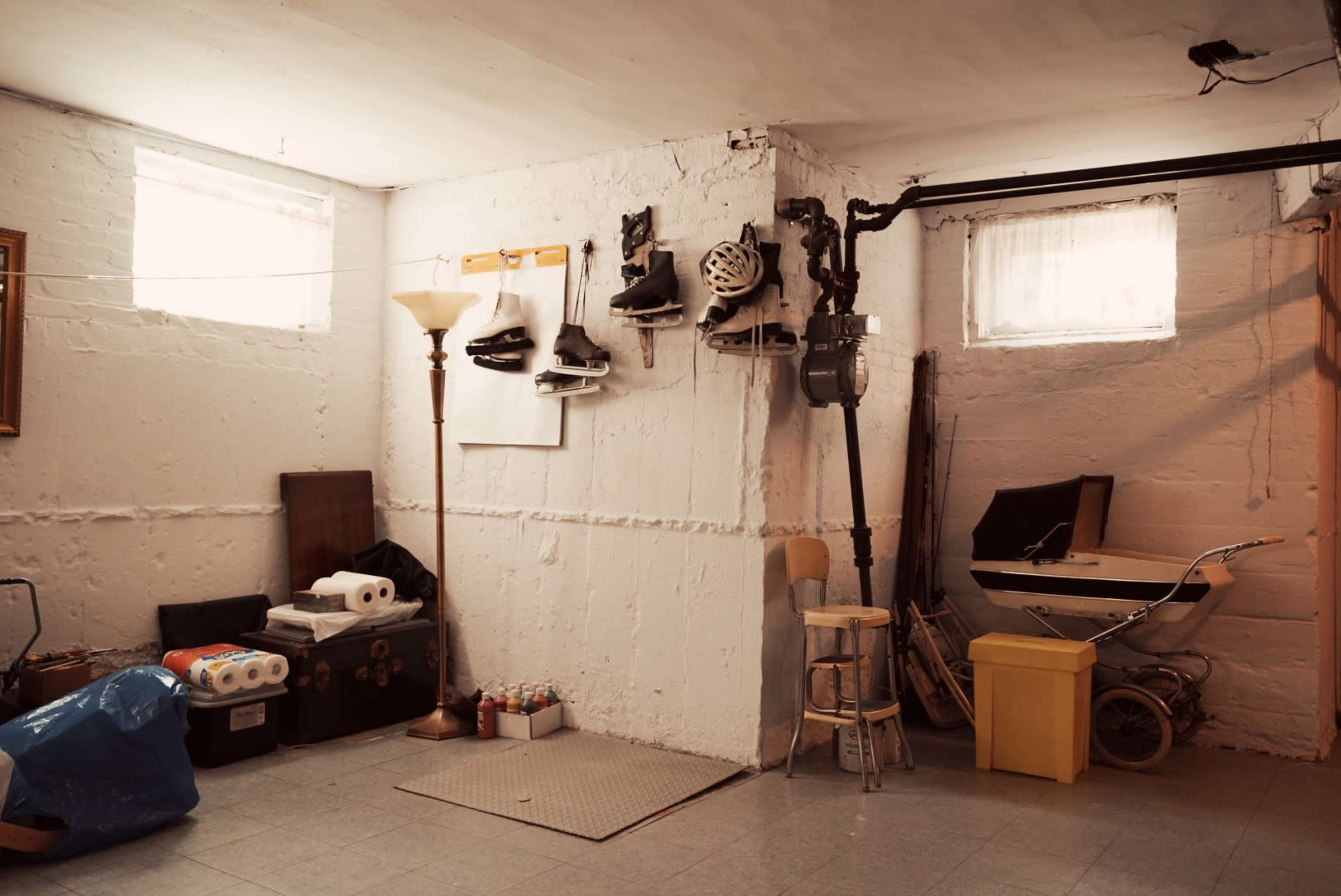 A basement room features a stack of boxes, a lamp, and various items including skates, a stroller, and rolls of paper against a white, unfinished wall.
