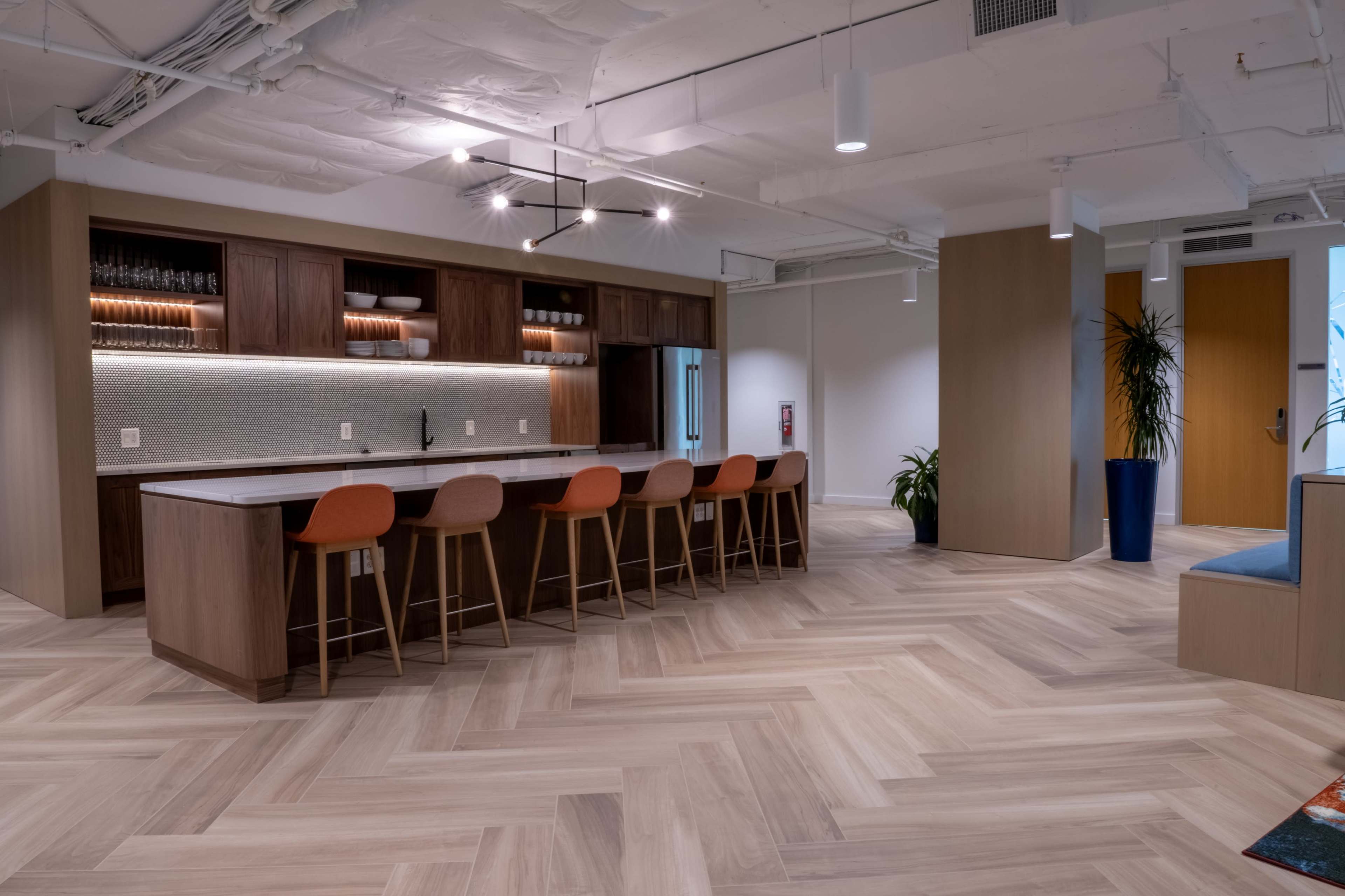 A modern office kitchen area featuring a long countertop with bar stools and wooden cabinetry, set against a backdrop of herringbone-patterned wood flooring.