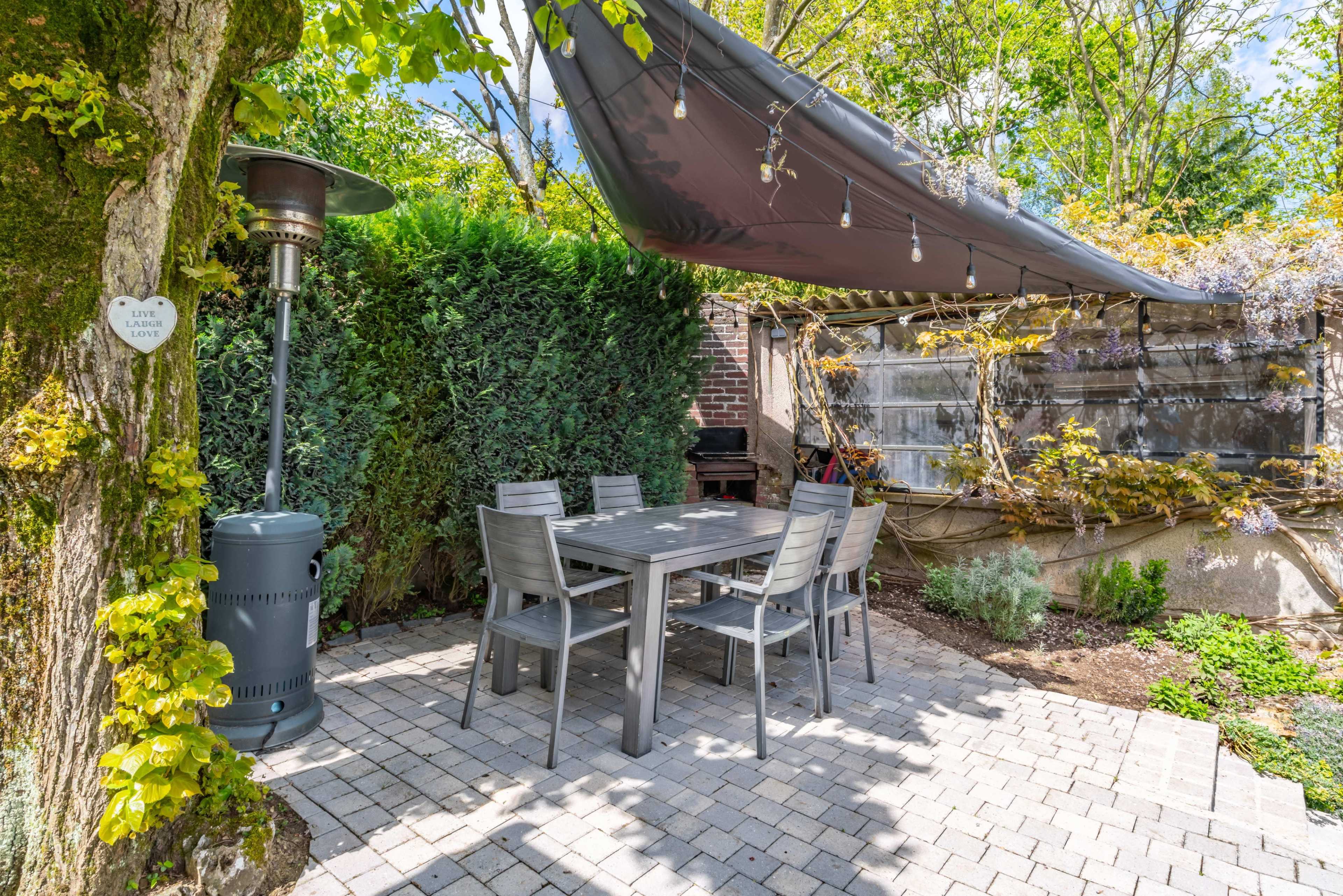 A patio area with a gray table and chairs, surrounded by greenery and a black canopy overhead.