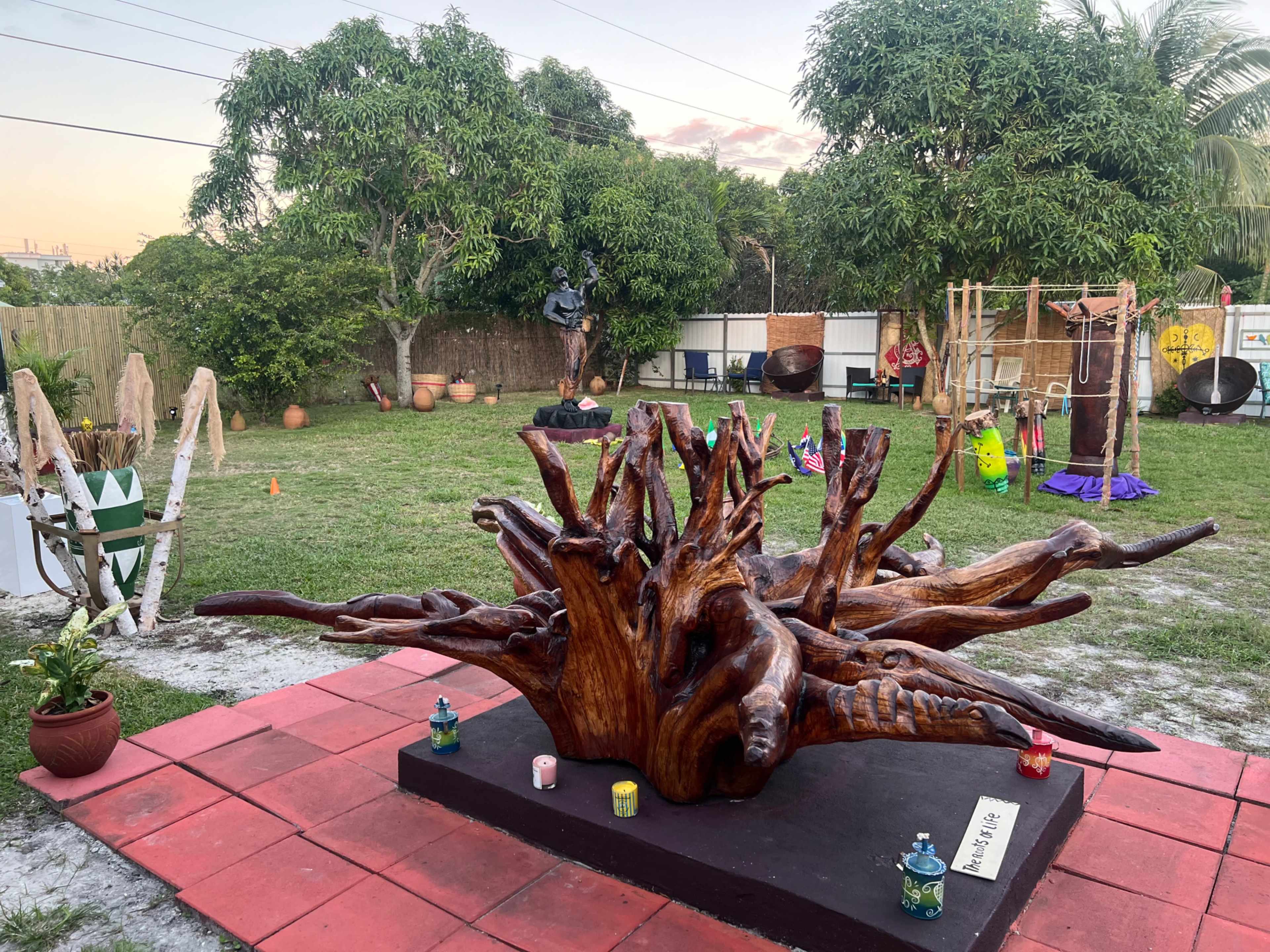 A grassy outdoor space featuring a large wooden sculpture at the forefront, surrounded by various decorations and plants, with trees and a play structure in the background.