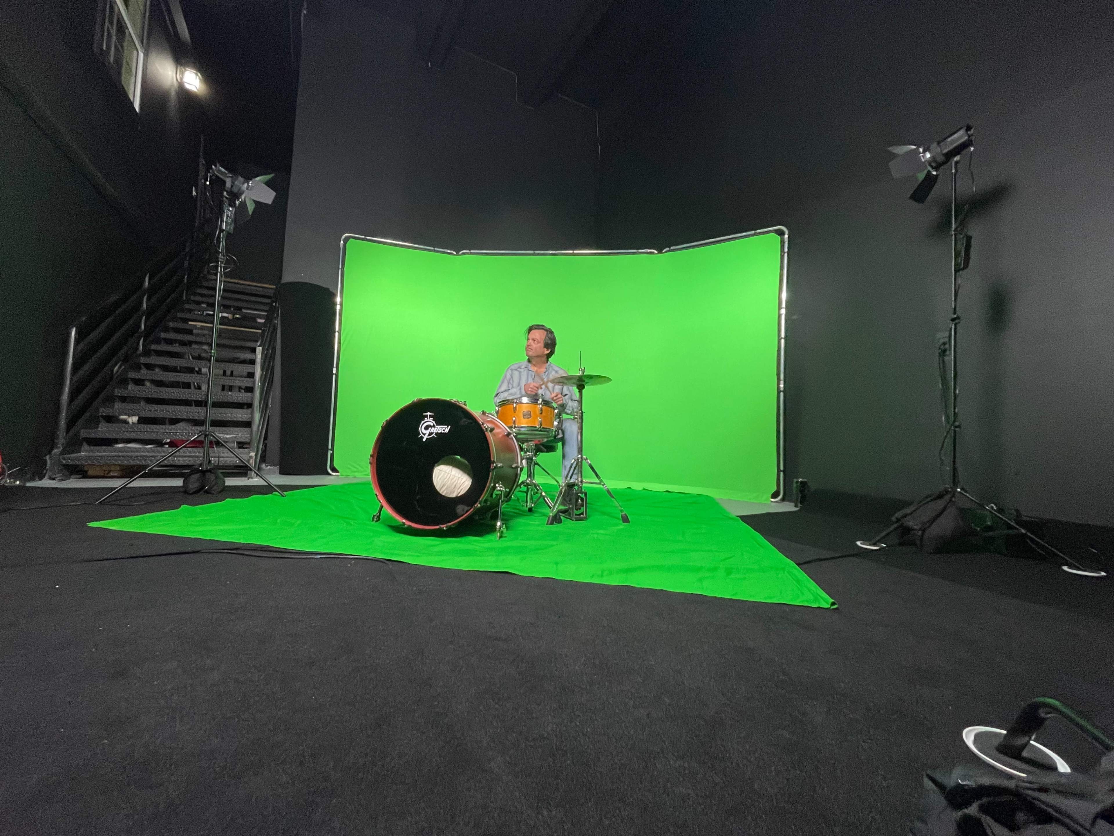 A drummer sits on a green screen set in a studio with lighting equipment and a staircase in the background.