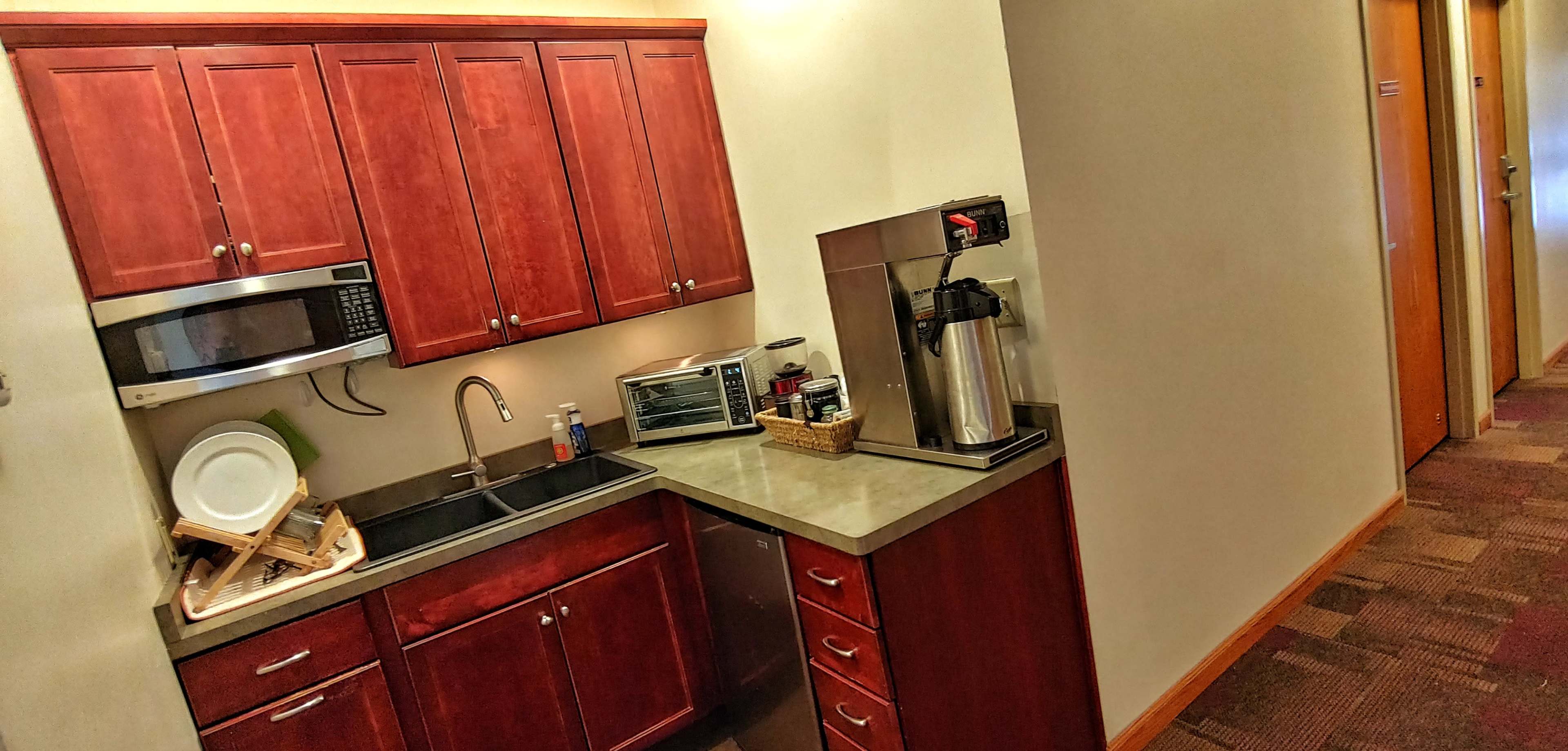 A kitchenette with wooden cabinets, a microwave, a sink, a countertop, and a coffee maker, adjacent to a hallway.