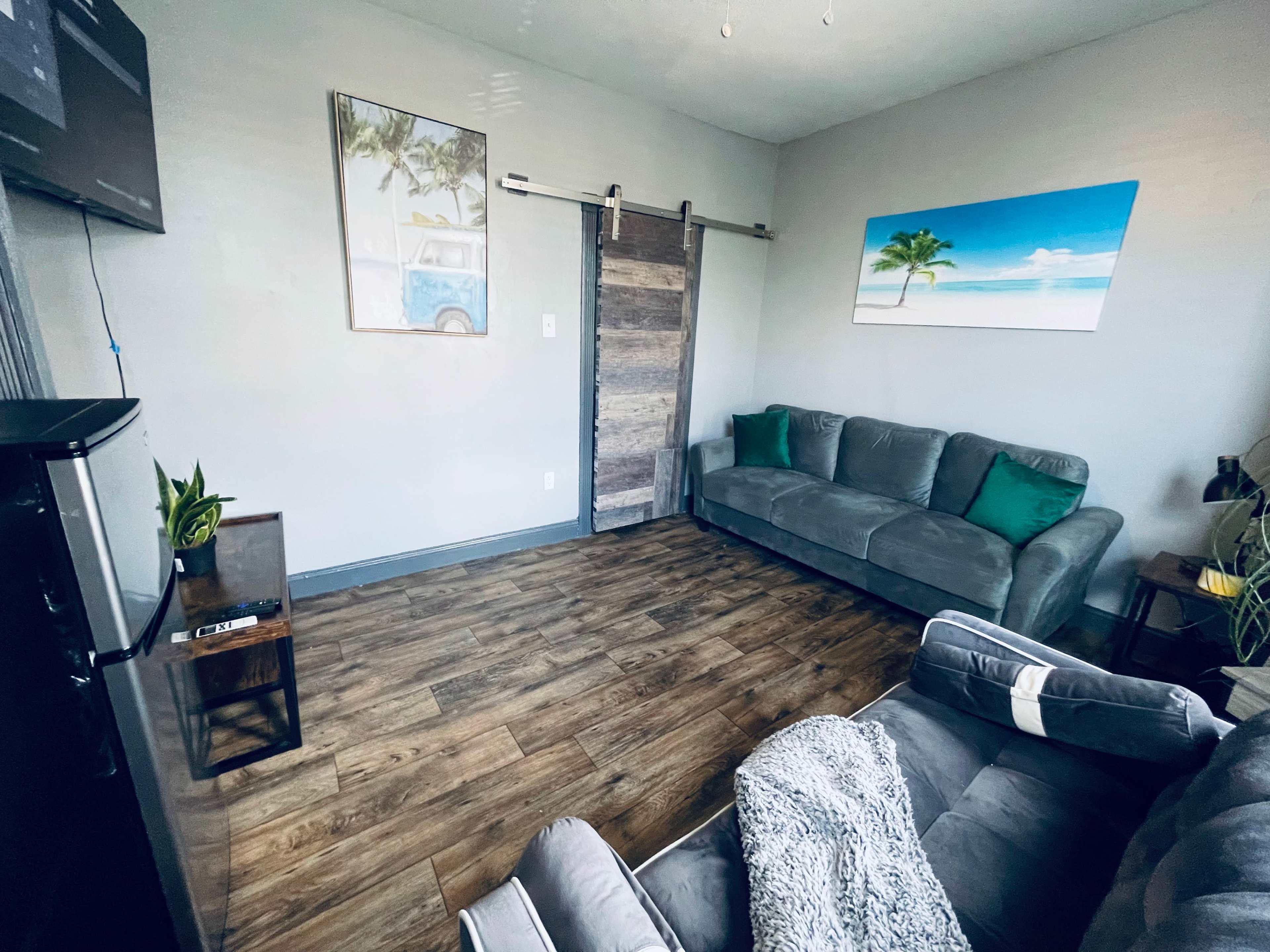 The room features a gray sofa, a sliding wooden door, and a mix of wall art depicting a beach scene and a vintage vehicle.