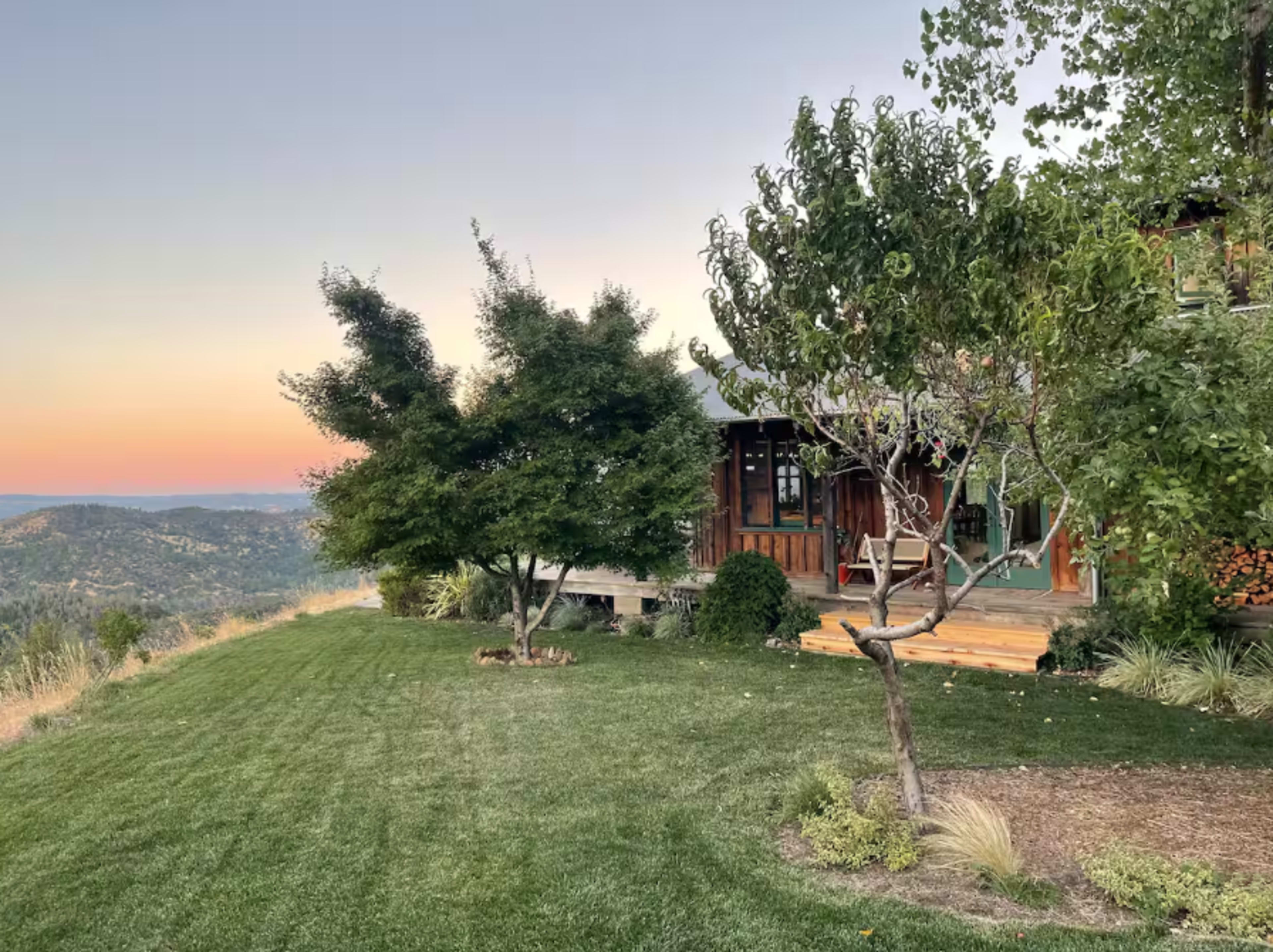 A wooden house sits on a hillside with two trees and a manicured lawn overlooking a distant valley at sunset.