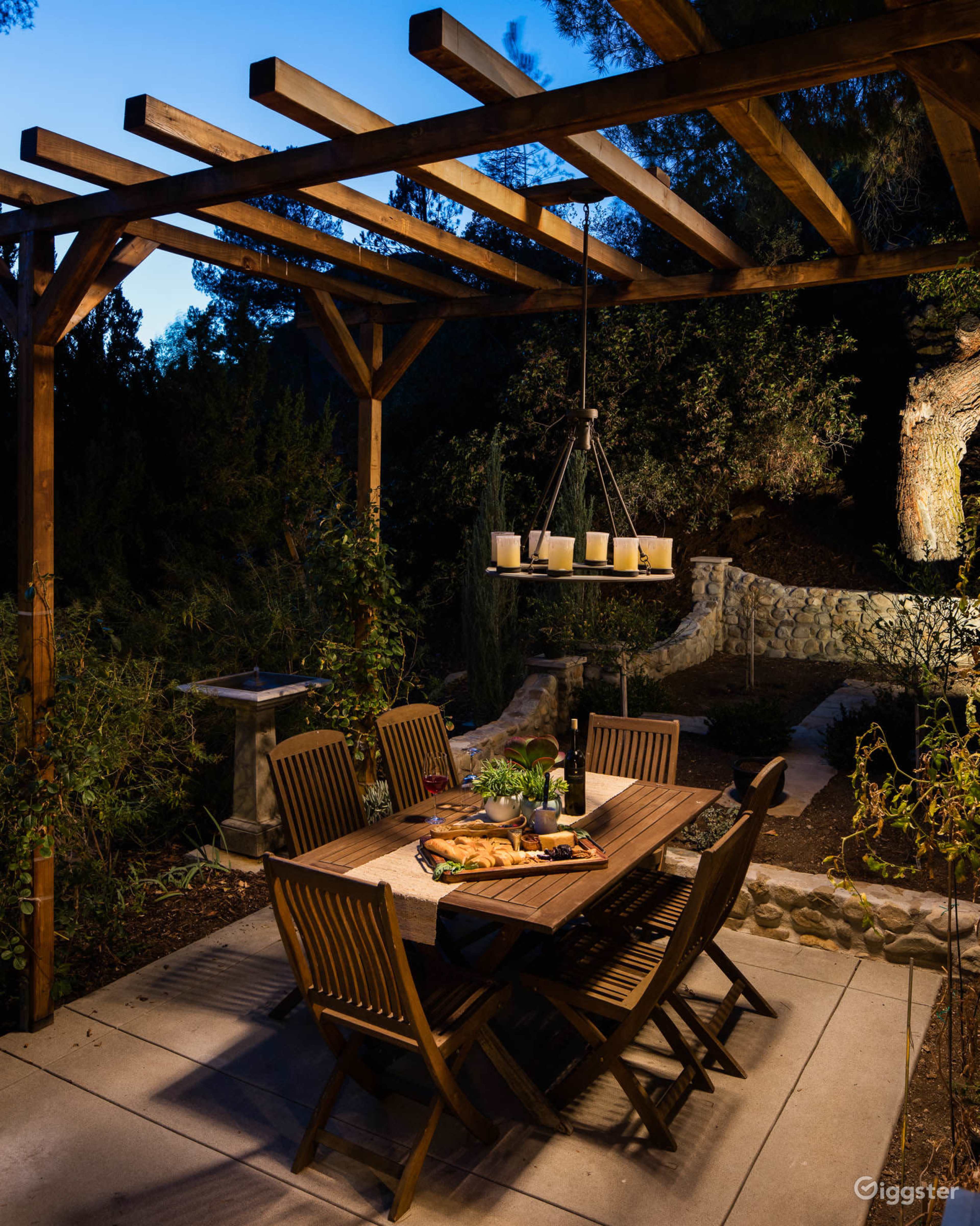 A wooden dining table is set with food and drinks under a pergola in an outdoor garden at dusk.