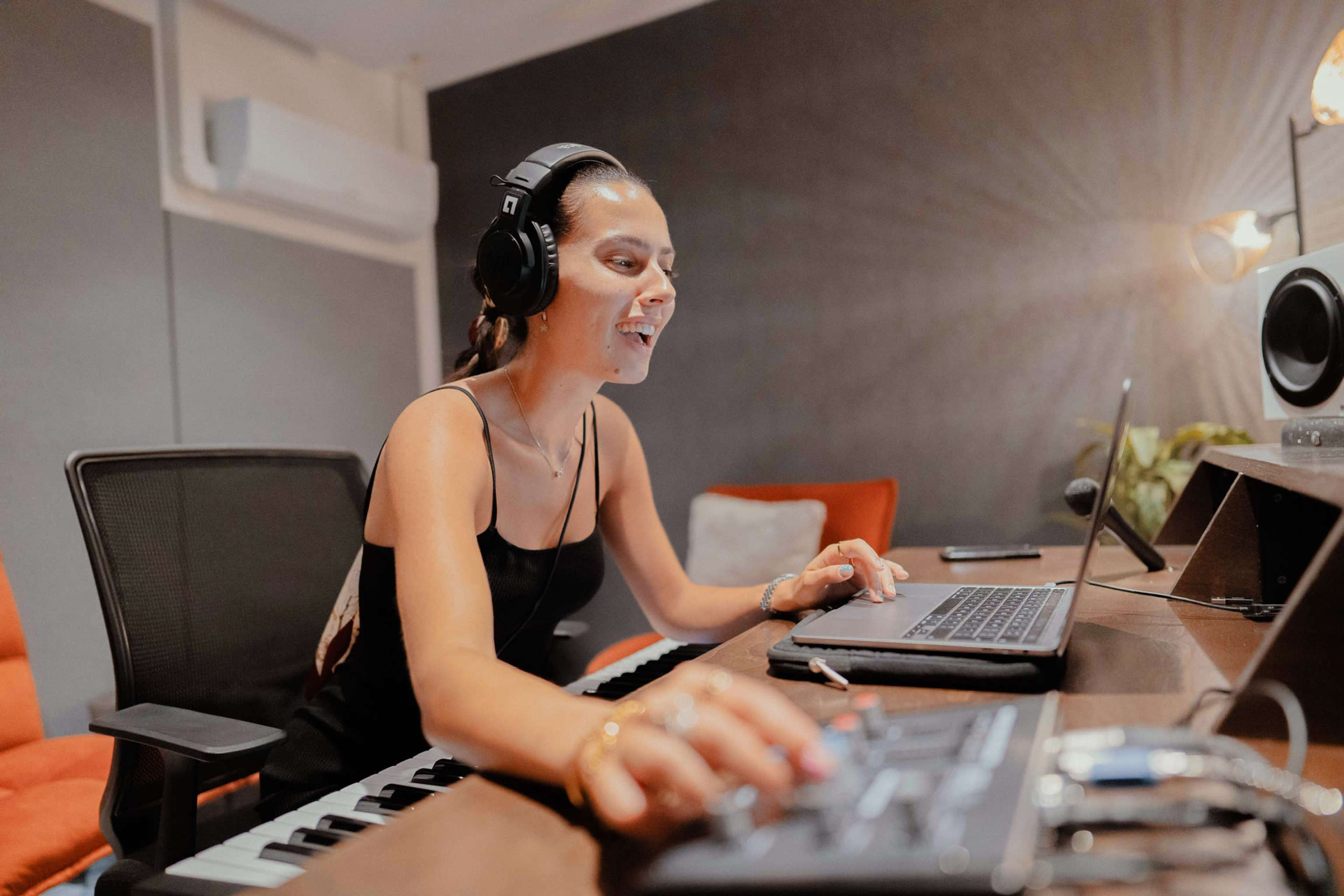 A woman with headphones sits at a desk, working on a laptop while using a keyboard in a music studio.