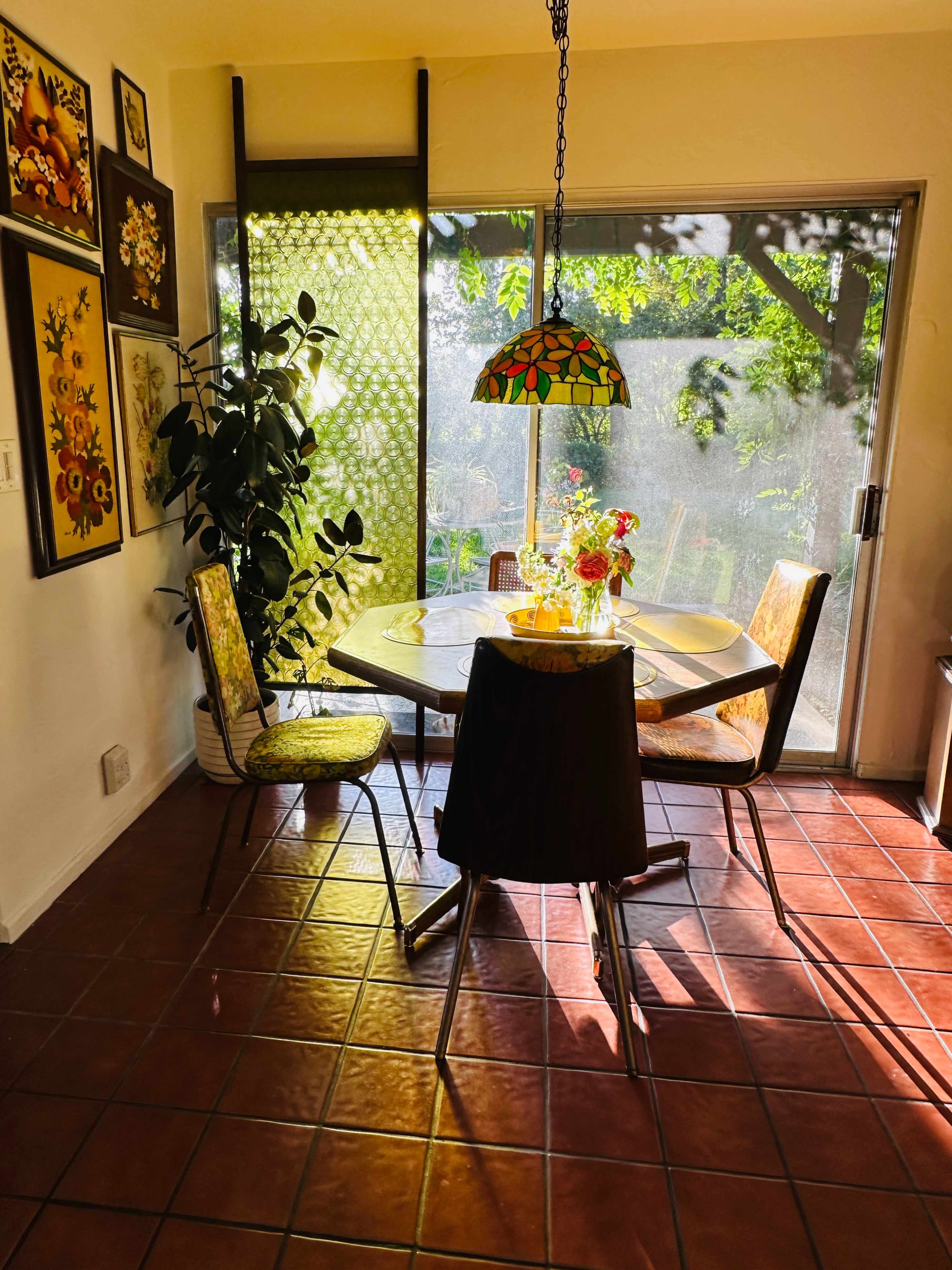A round dining table with a stained glass lamp is surrounded by four chairs in a brightly lit kitchen near a window overlooking greenery.