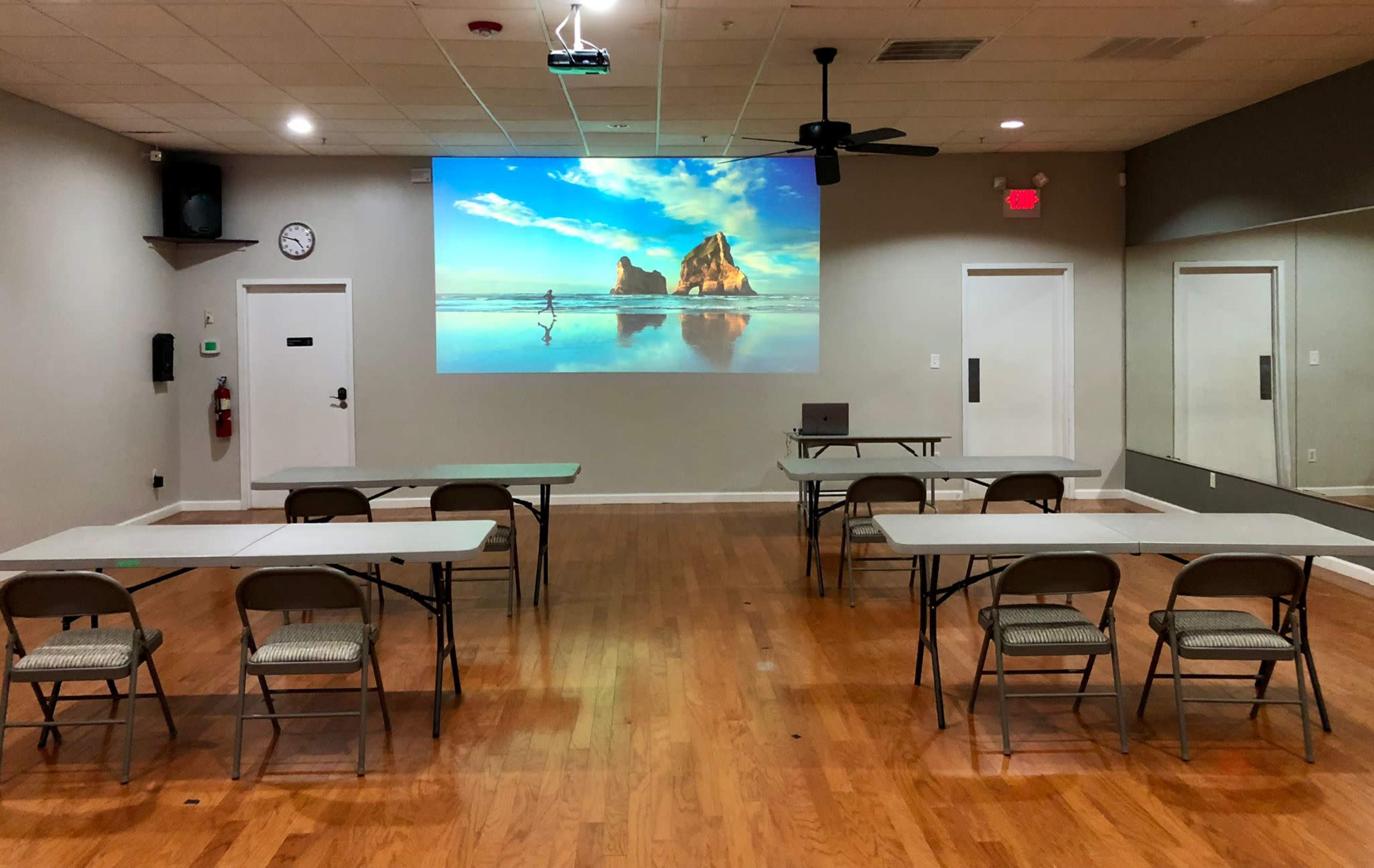 A meeting room features several tables and chairs arranged facing a large projection screen displaying a beach scene.