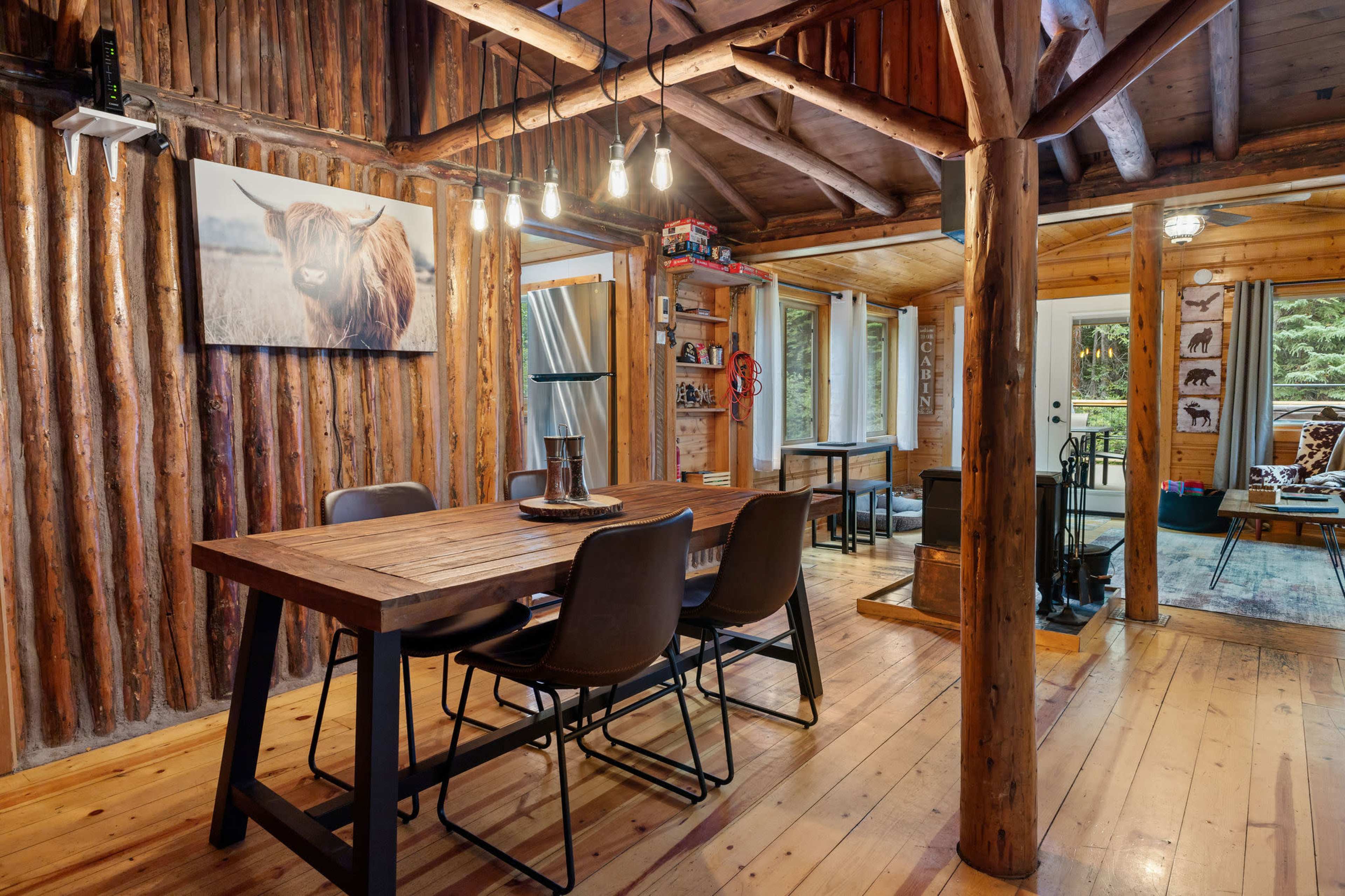 The image shows a rustic dining area with a wooden table and chairs, surrounded by log walls and a cabin-style interior, featuring a wall-mounted artwork of a cow.