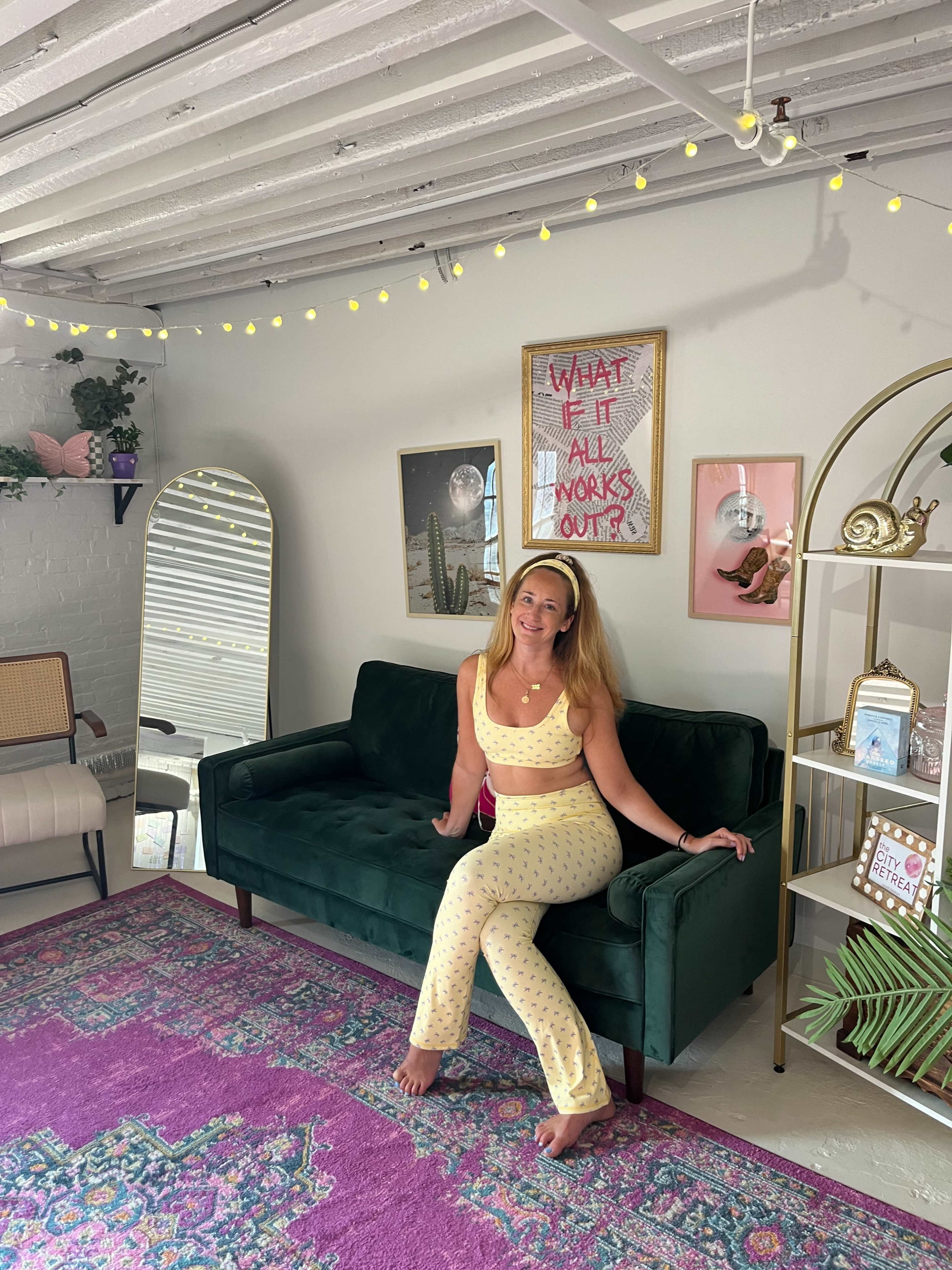 A woman sits on a green couch in a brightly decorated room featuring a mirror, colorful artwork, and a vibrant rug.