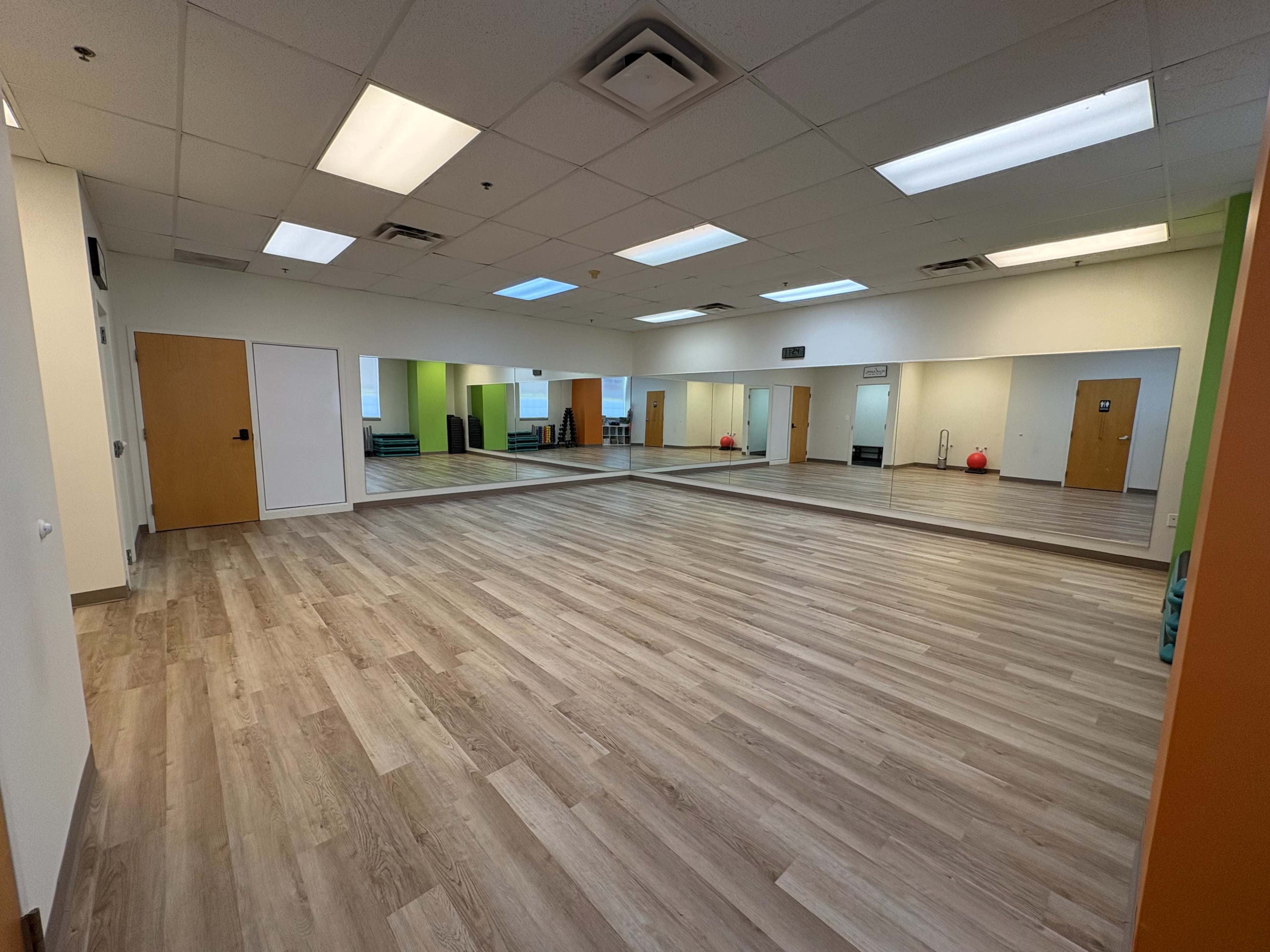 The image shows a spacious, well-lit room with wooden flooring and large mirrors on one wall, along with a door and colorful accents.