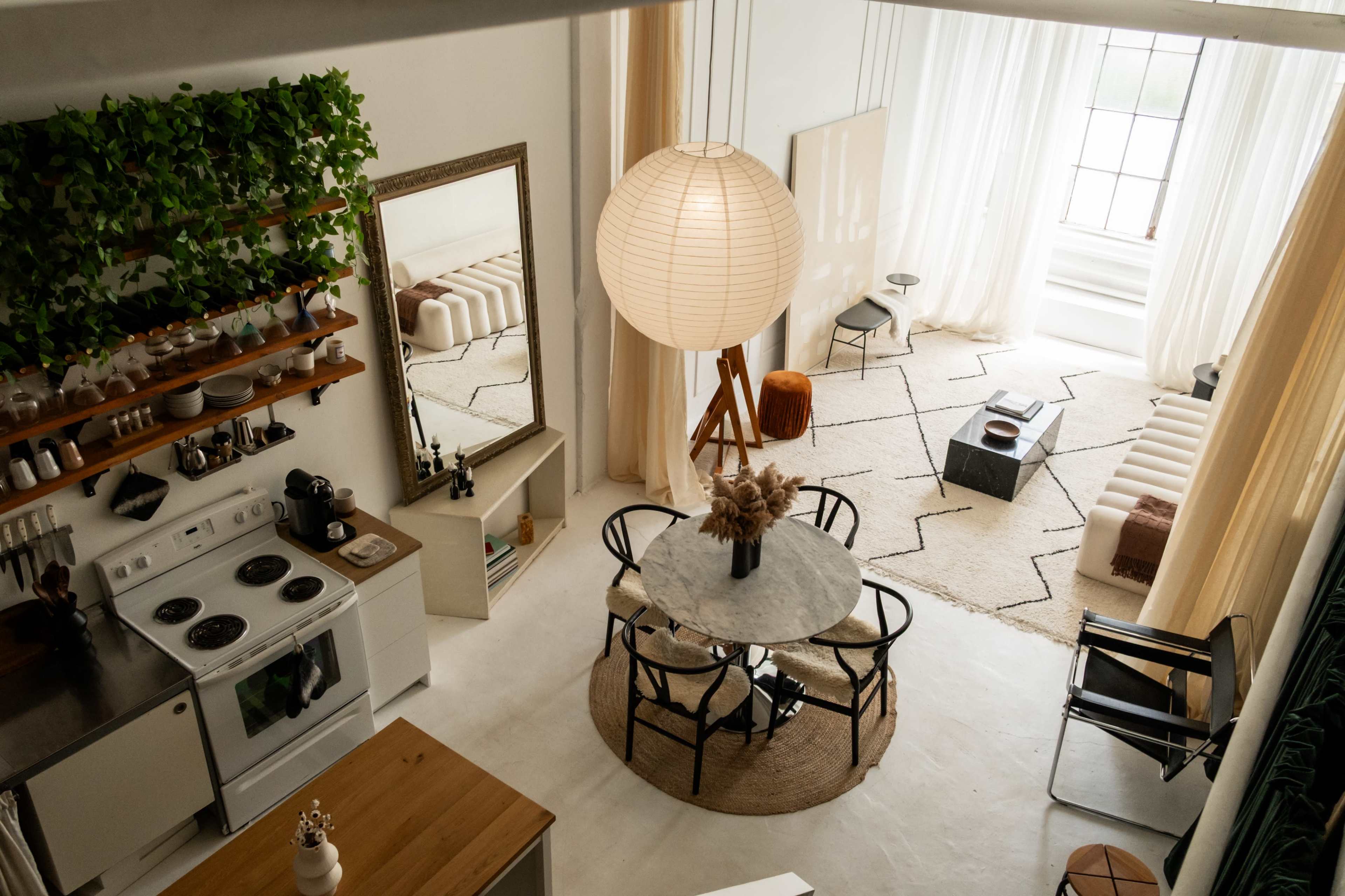 The image showcases a stylish, open-concept living space featuring a kitchen, dining area with a round table, and a cozy lounge area with large windows and soft furnishings.