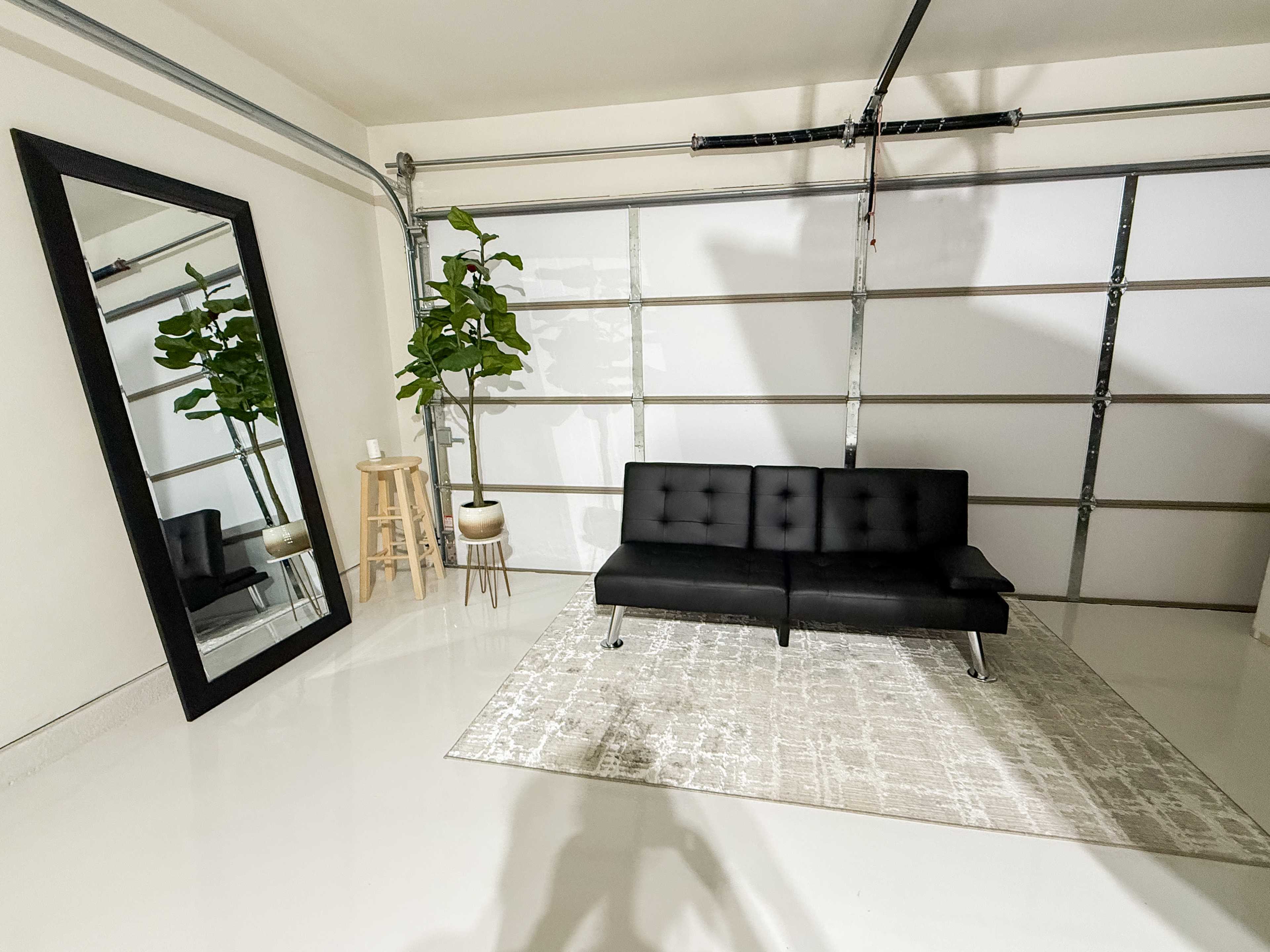 The image shows a minimalist garage space featuring a black sofa, a large mirror, a potted plant, and a rug on a white floor.