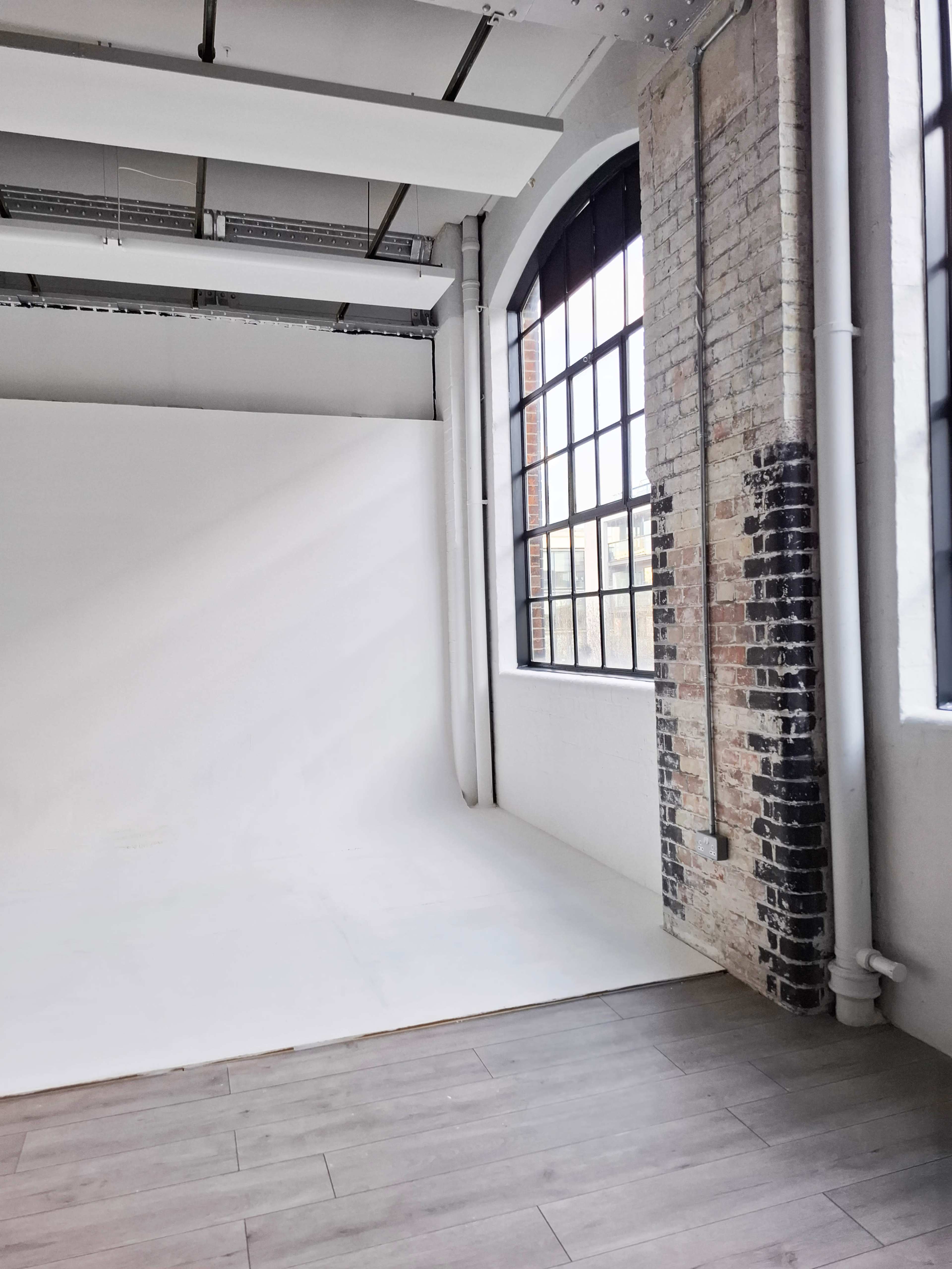 The image shows a spacious interior with large windows, exposing a white backdrop and brick walls in a setting that appears to be a photography studio.