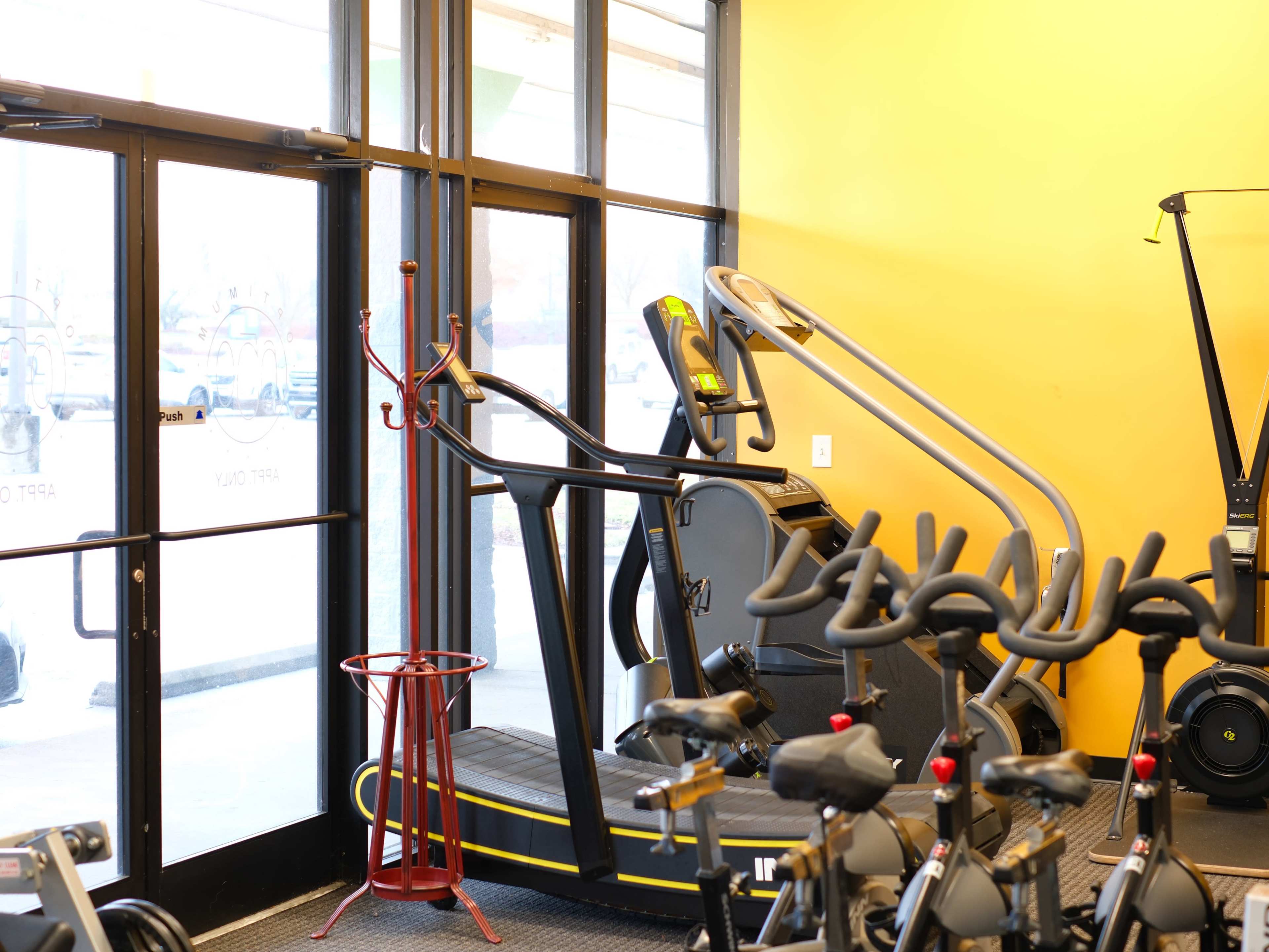 The image shows a fitness center interior with exercise equipment, including an elliptical and a treadmill, near a glass entrance.